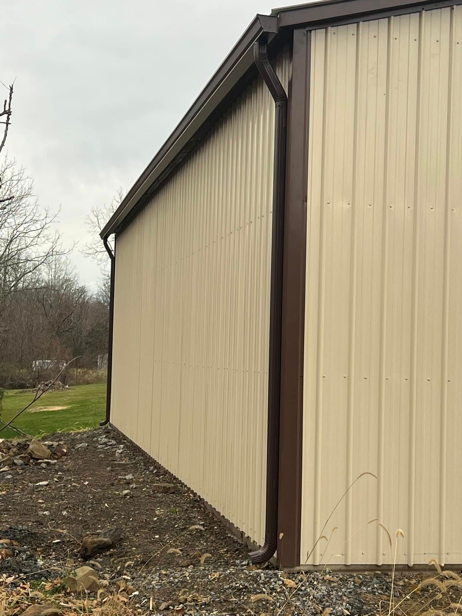 The corner of a metal building with tan siding, dark brown trim, and a matching downspout leading to gravel ground.