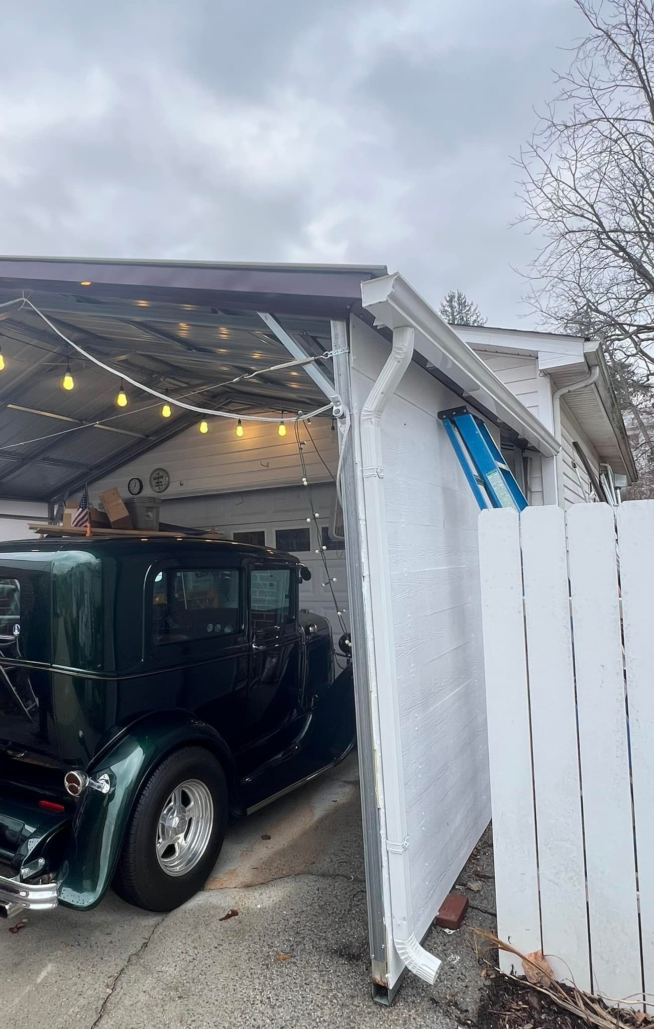 A vintage dark green car parked under a white carport with string lights, next to a white fence.