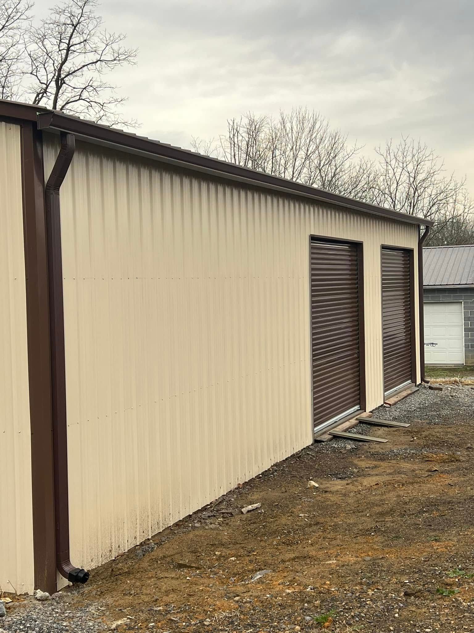 A side view of a cream-colored metal building featuring two brown rolling garage doors and a brown gutter downspout.