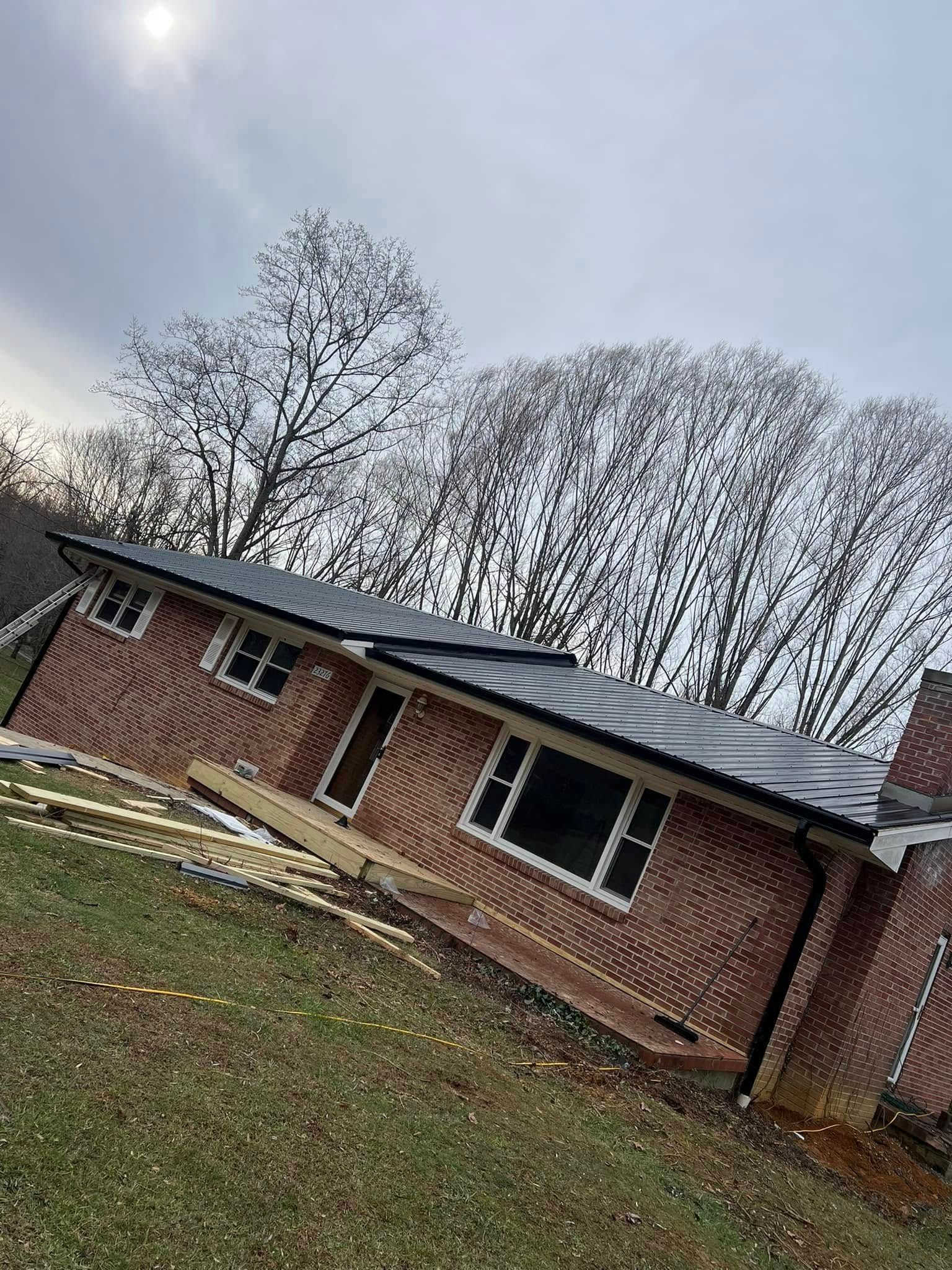 A brick ranch-style house with a dark metal roof, set against a backdrop of leafless trees under a cloudy sky.