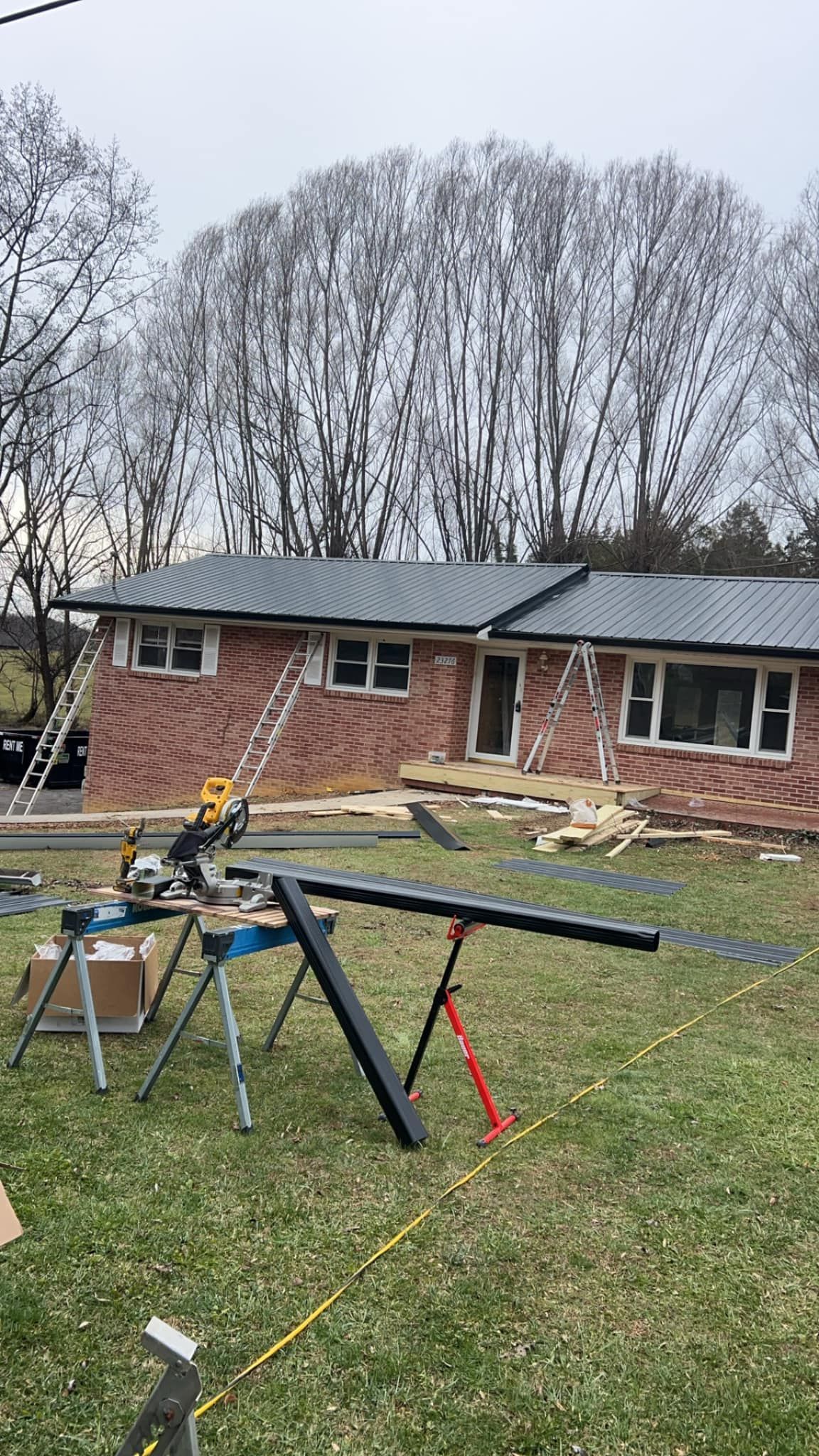 A brick house with a new dark metal roof undergoing renovations, featuring construction tools and equipment in the yard.