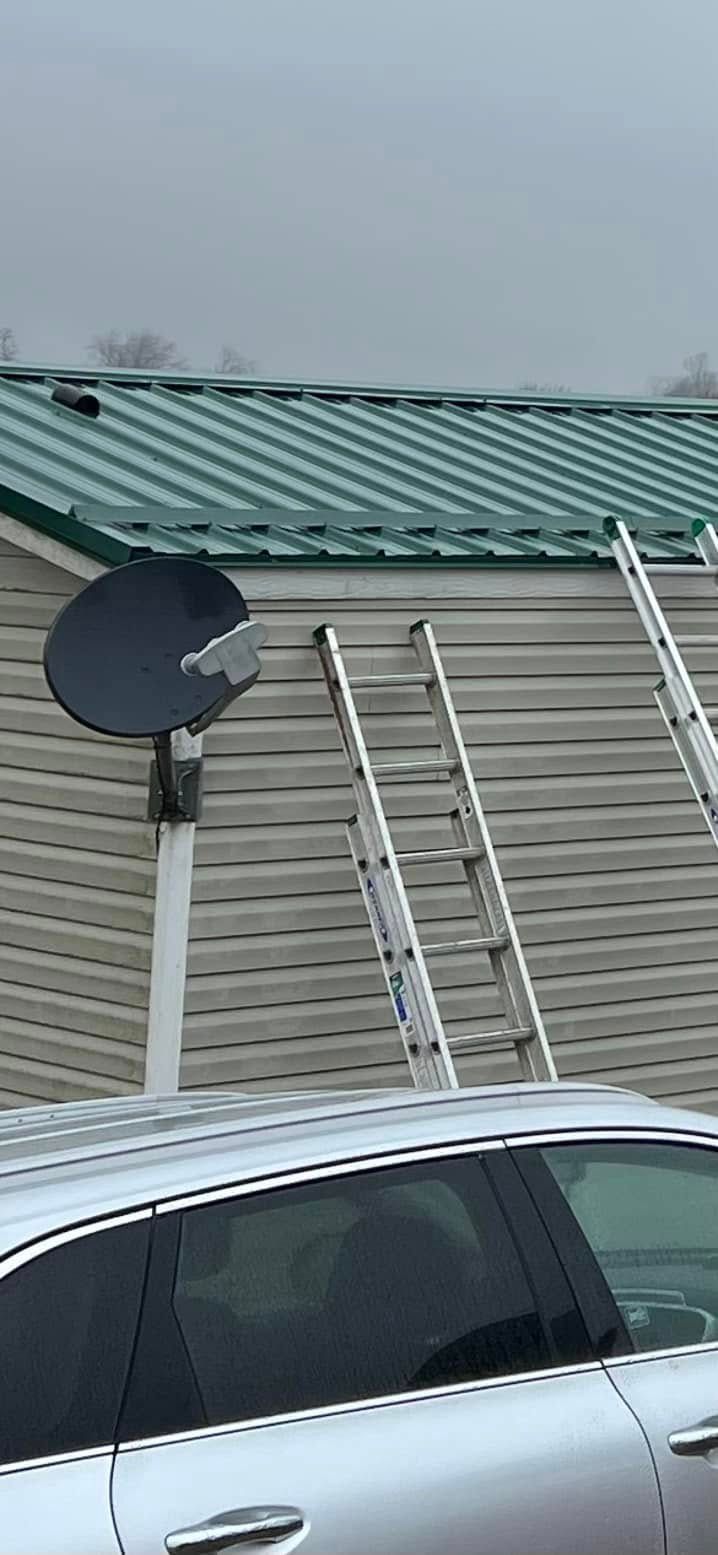 Two extension ladders lean against the horizontal beige siding of a building with a green metal roof under a gray sky.