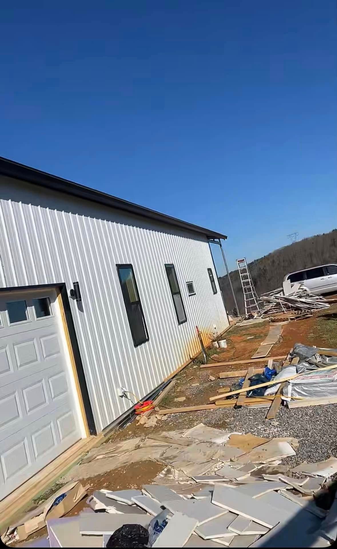 White metal-sided house with a garage and windows, with construction debris on the ground against a clear blue sky.