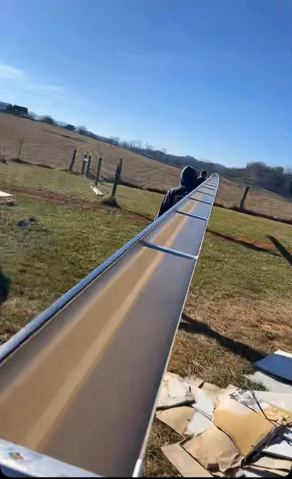 A long, metallic gutter segment resting on the ground in a grassy field with a person standing at the far end.
