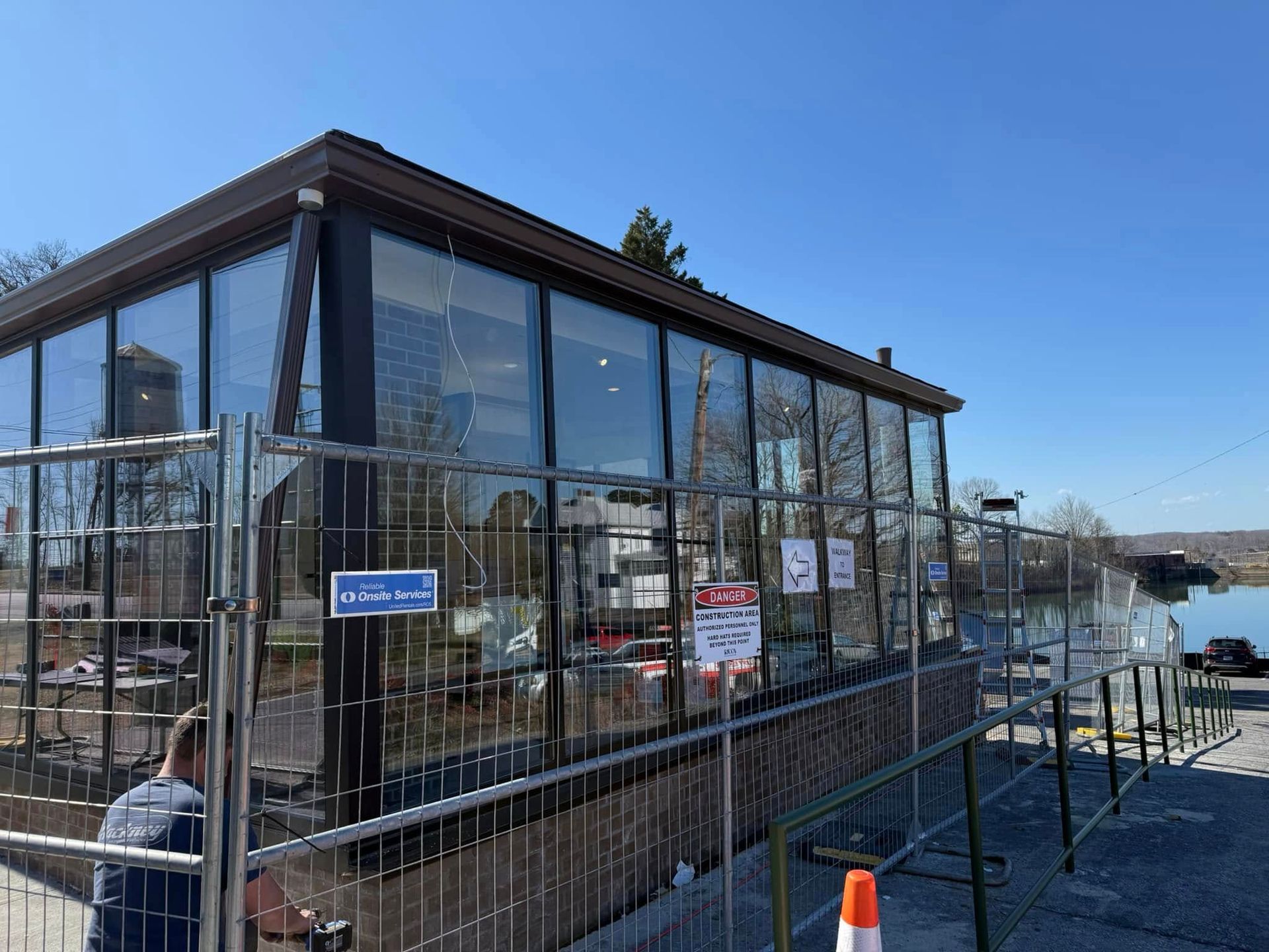 Modern glass building under construction with chain-link fencing, situated near a body of water under a clear blue sky.