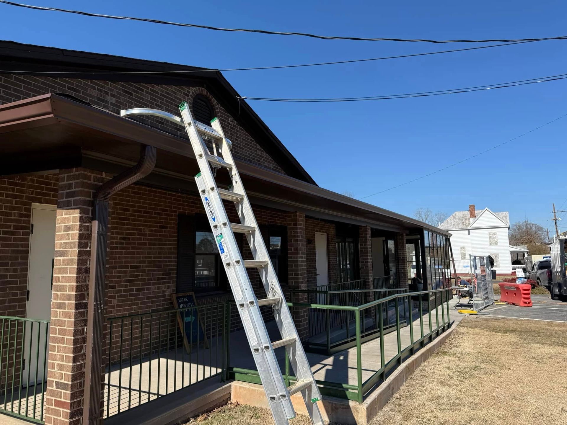 A metal extension ladder leans against the roofline of a brick building with a porch, under a clear blue sky.