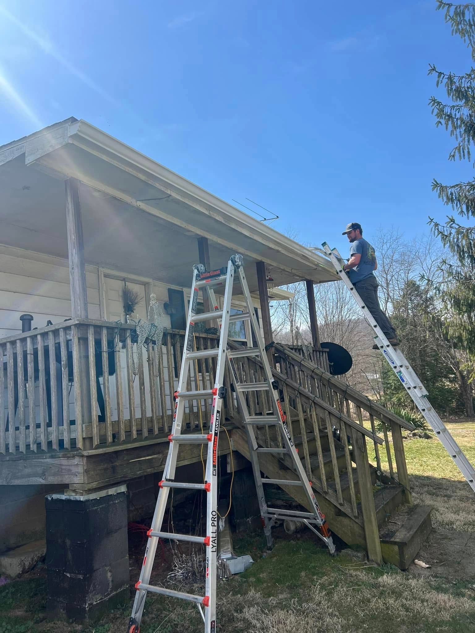 A person stands on a ladder against the eaves of a porch, while another ladder leans against the deck stairs nearby.