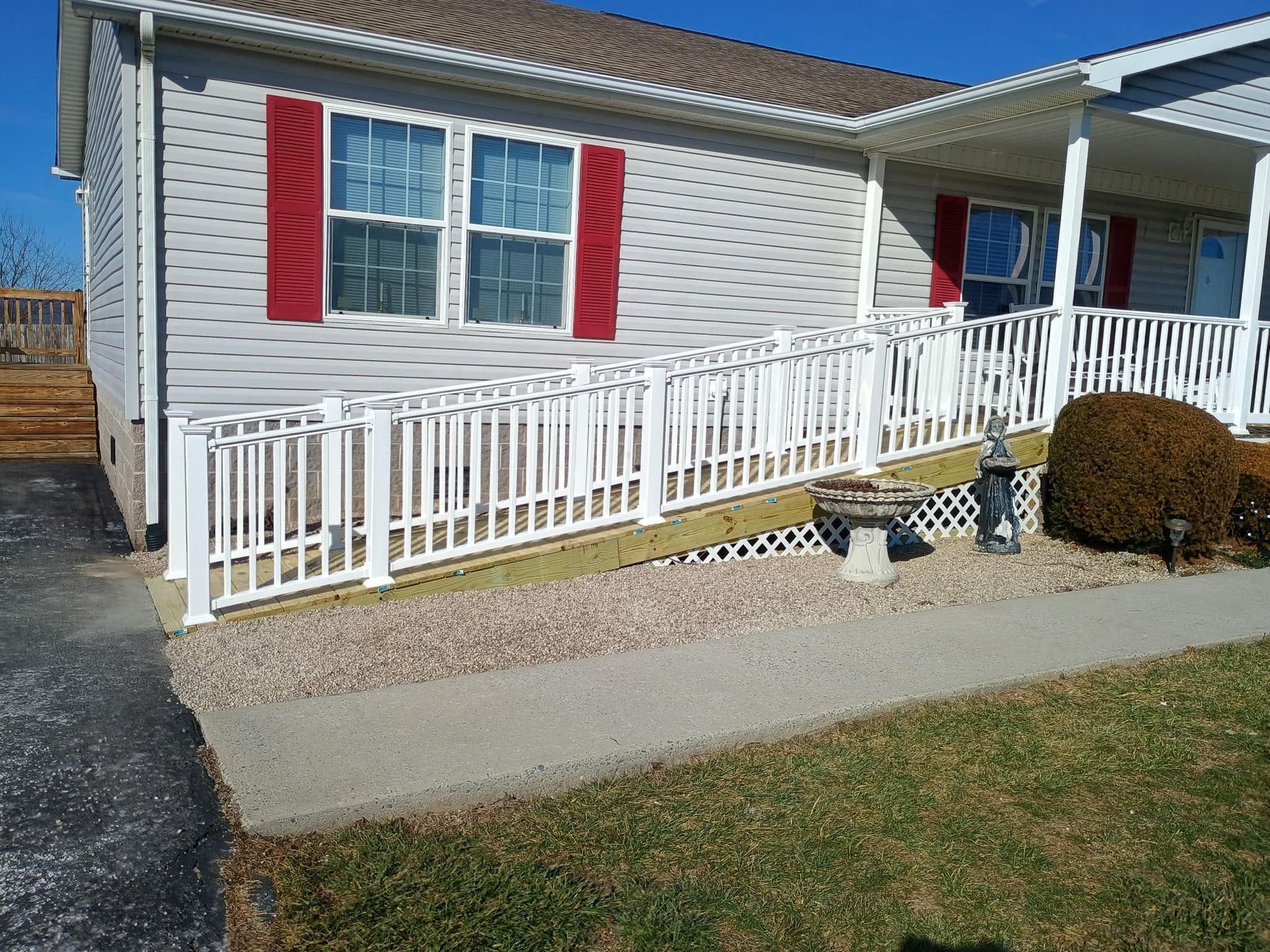 A white wheelchair ramp with matching railings leads up to the front entrance of a grey house with red window shutters.