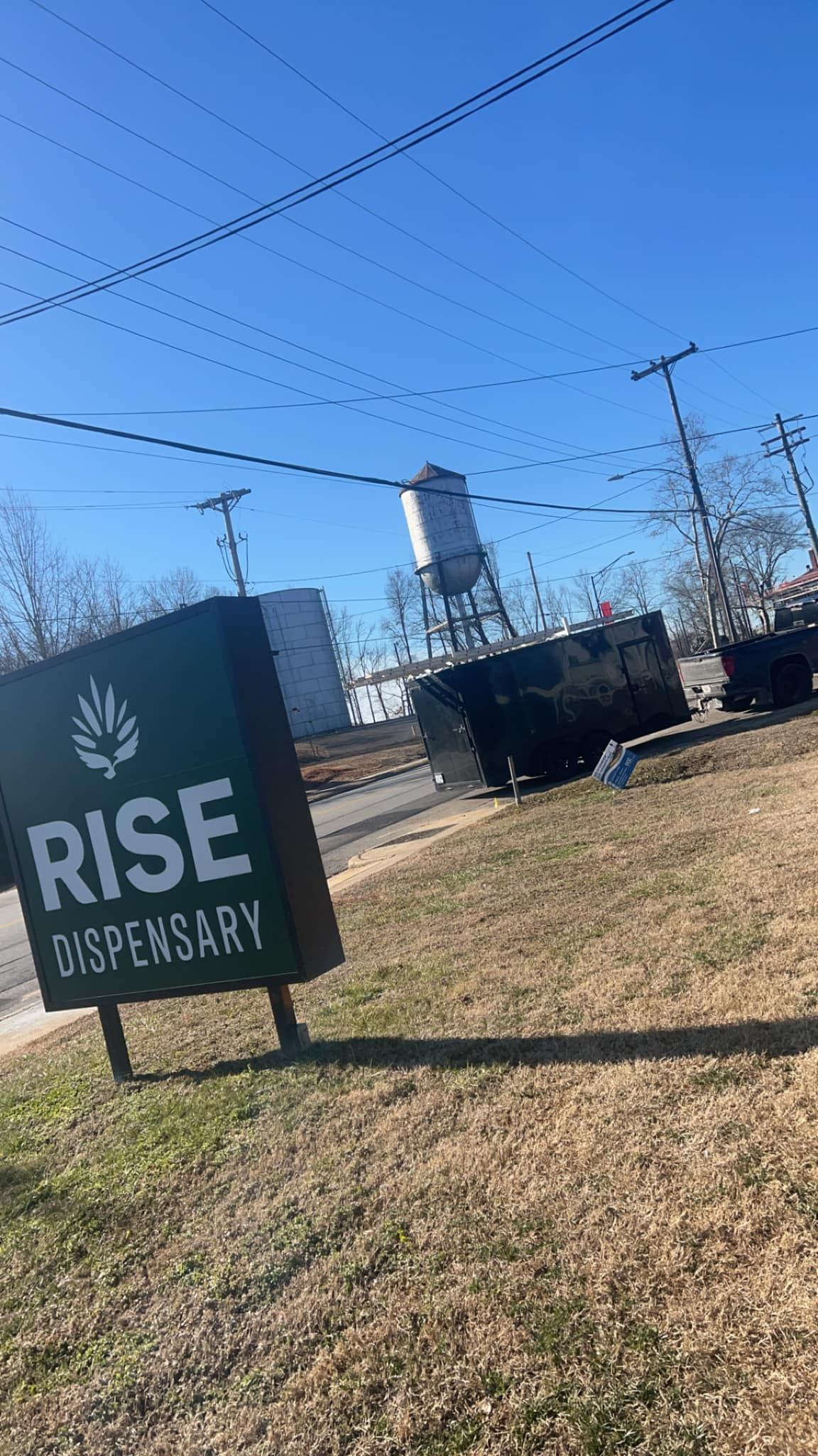 A green Rise Dispensary sign stands on a grassy lawn under a clear blue sky, with a water tower visible in the background.