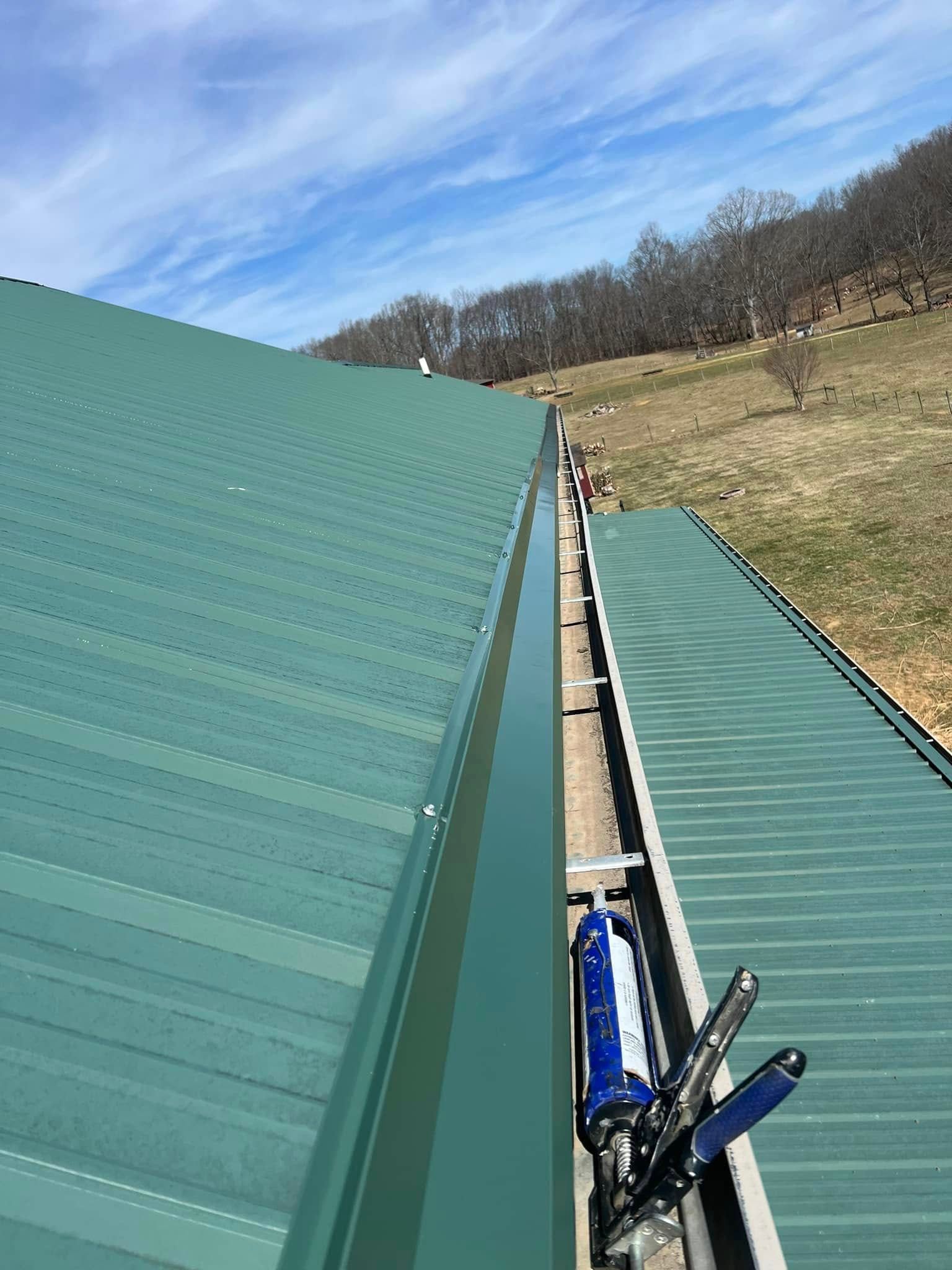A caulk gun and pliers rest in the gutter between two sections of a green metal roof under a blue, cloudy sky.