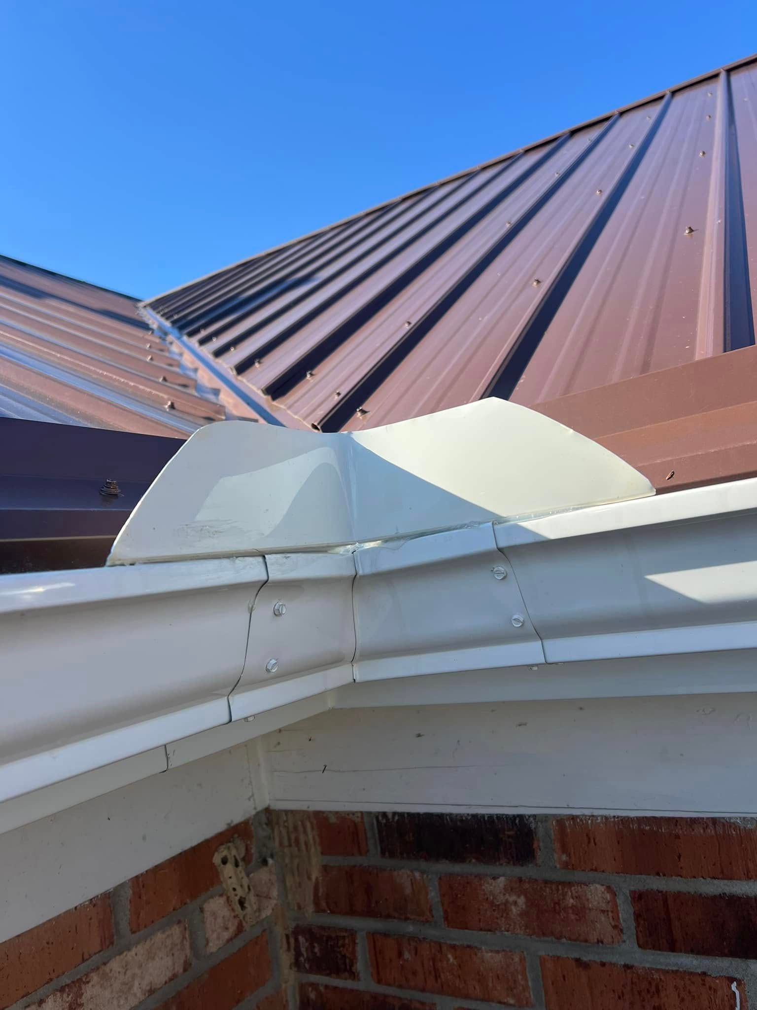 White metal gutter splash guard installed at a roof corner junction with a brown metal roof against a brick wall.