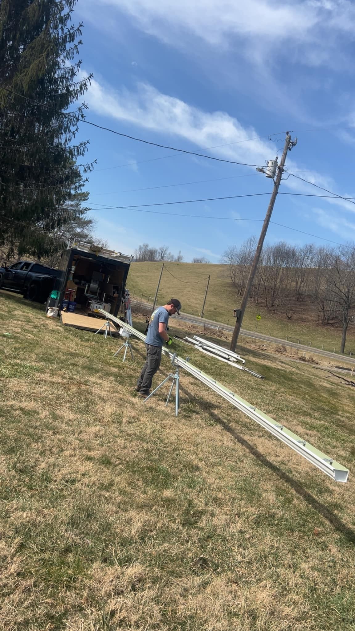 A person works on a long, metal lattice tower segment supported by a tripod in a grassy field near a utility pole.