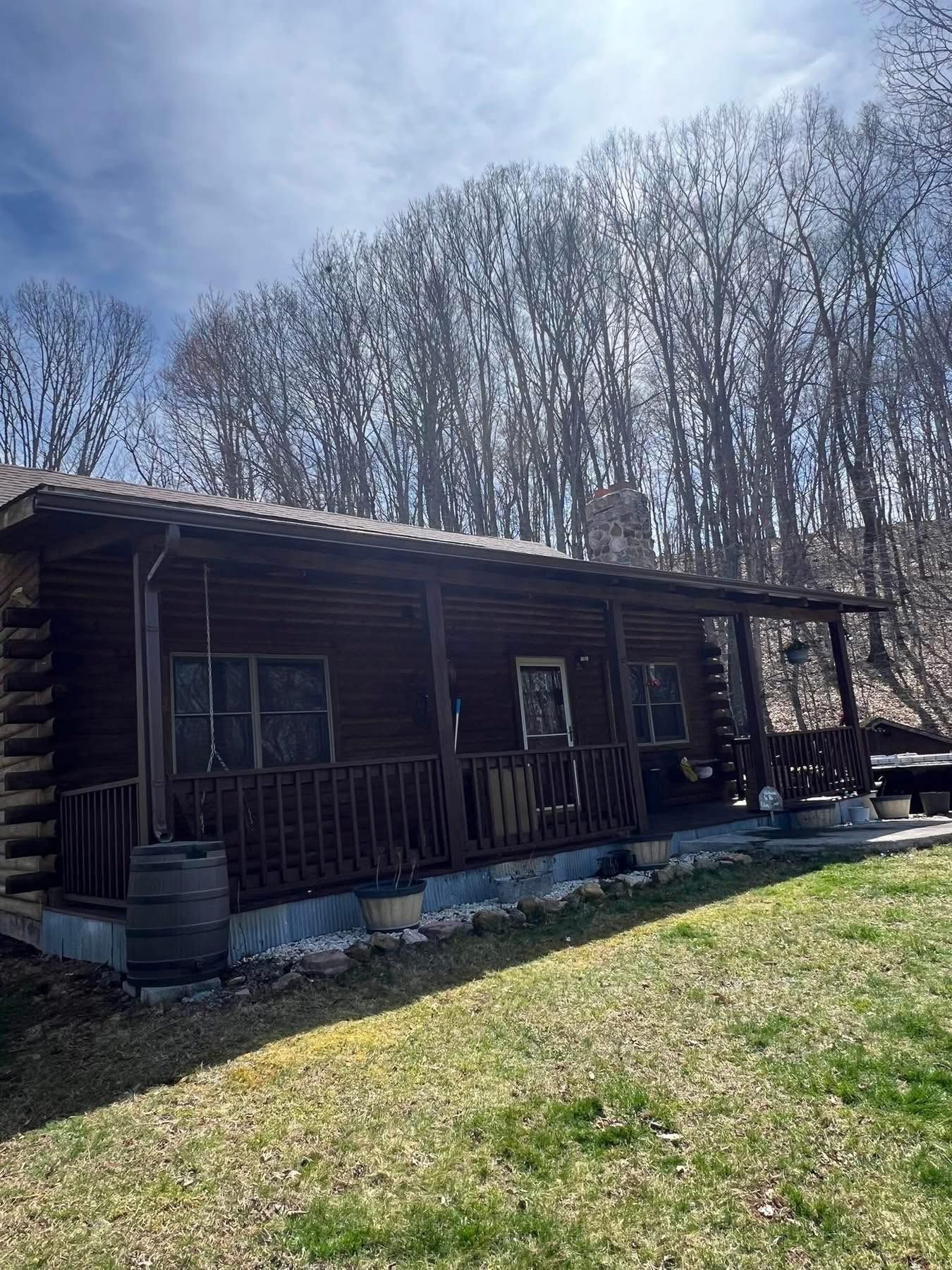 A rustic dark brown wooden log cabin with a covered front porch, situated in a wooded area under a blue sky.