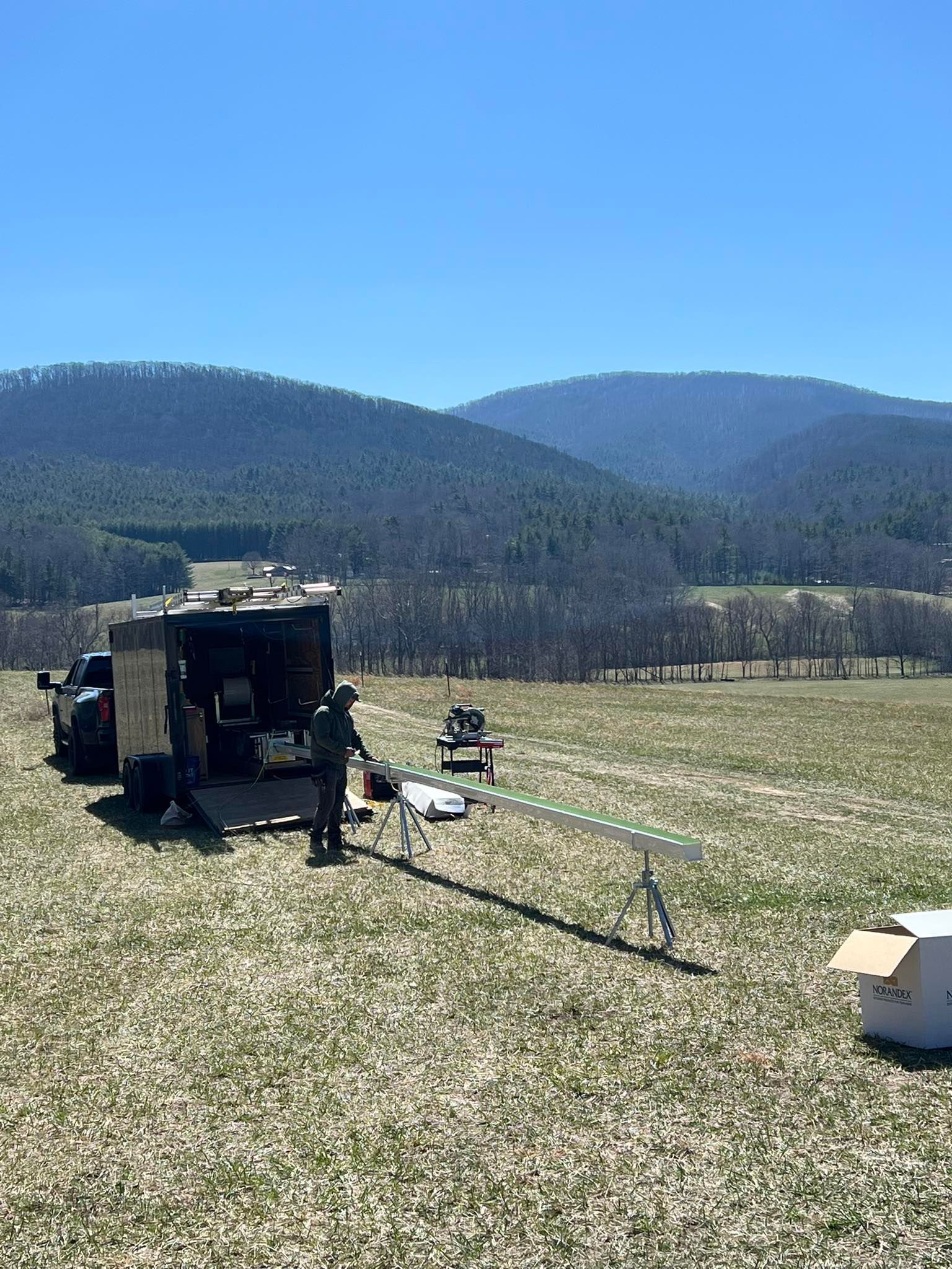 A person stands next to a mobile trailer and a long sensor arm set up in a field, with mountains in the background.