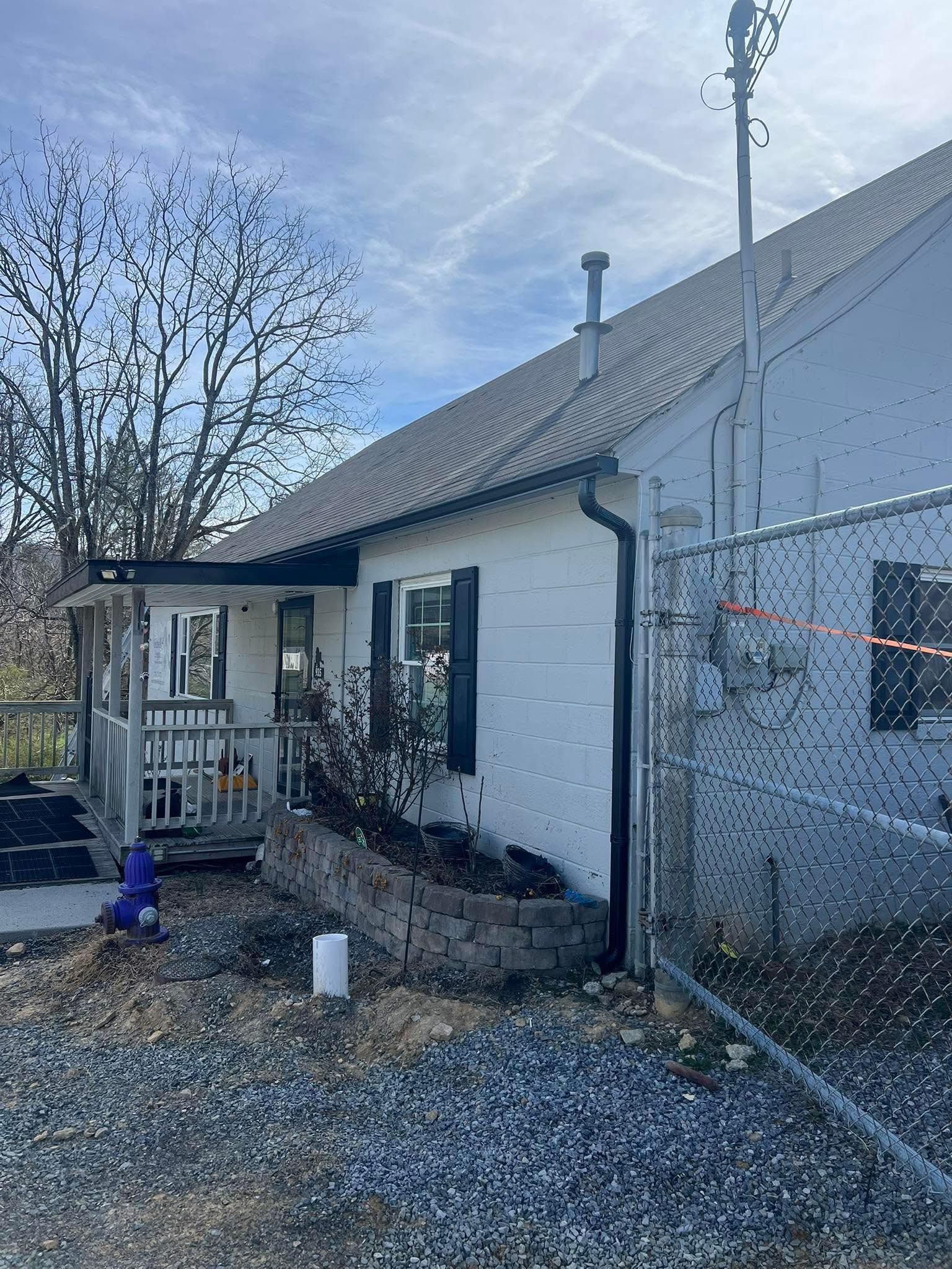 A white house with a black-roofed porch, stone planter, and a chain-link fence under a blue sky.