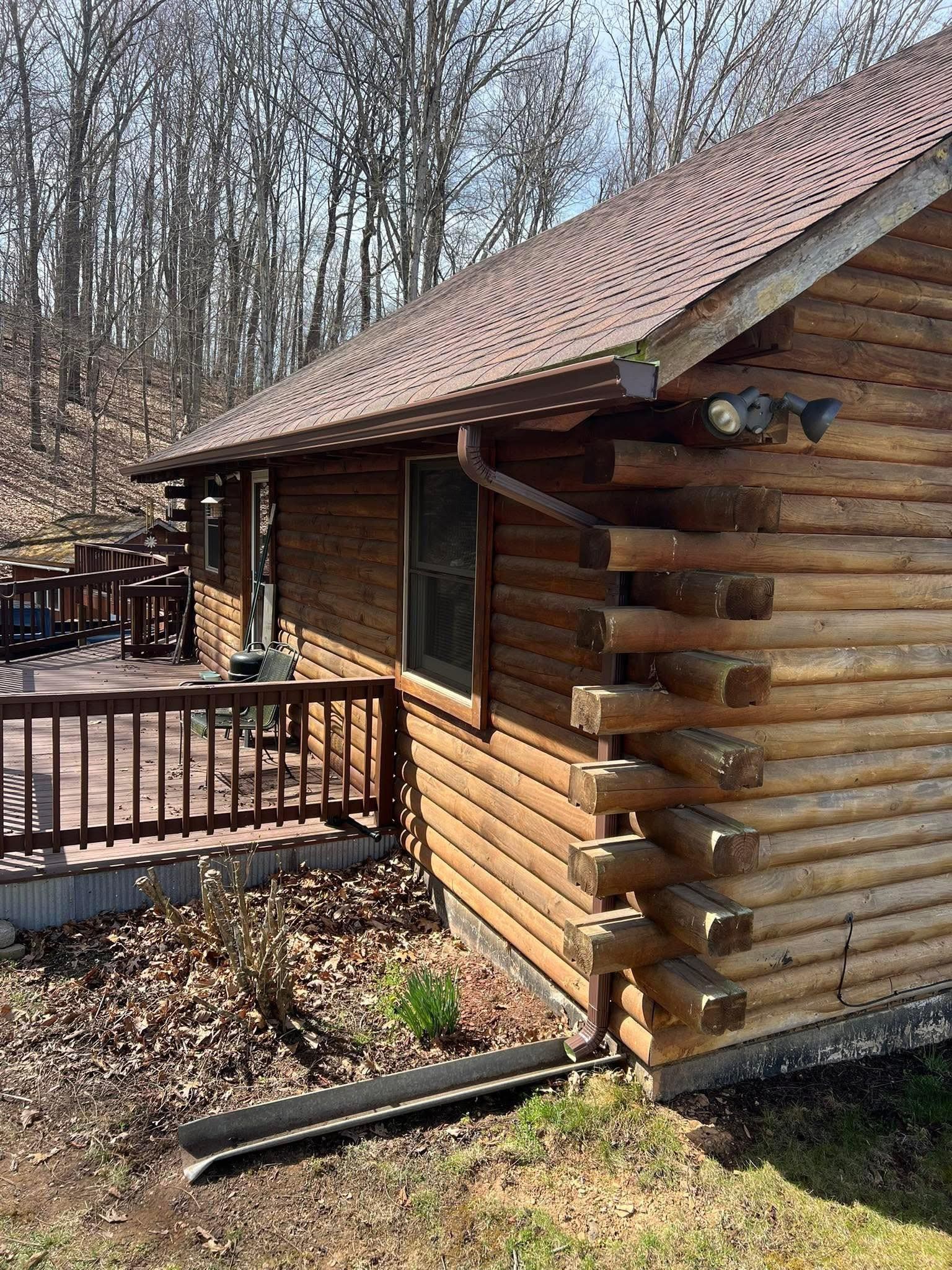 A brown log cabin sits beside a wooden deck, with a downspout extending into the yard against a backdrop of bare trees.