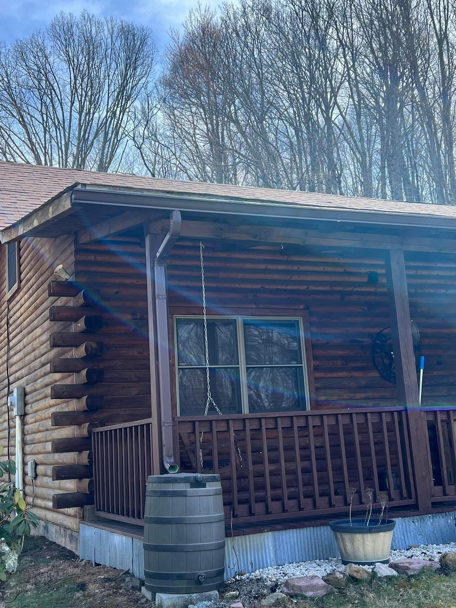 A brown log cabin with a porch, a metal roof, and a rain barrel collecting water from the gutter downspout.