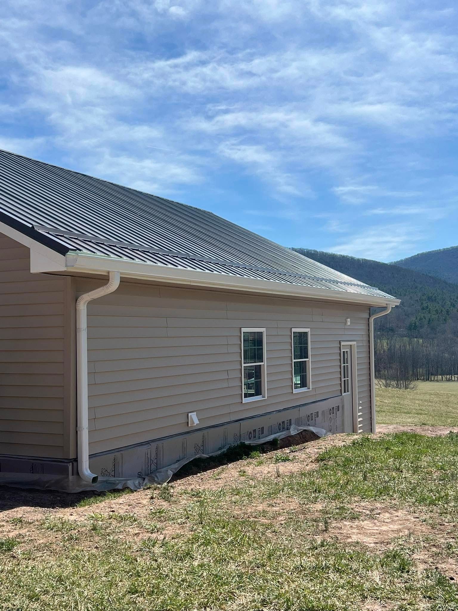 A tan, single-story house under construction with a dark metal roof, set in a field against a backdrop of distant hills.