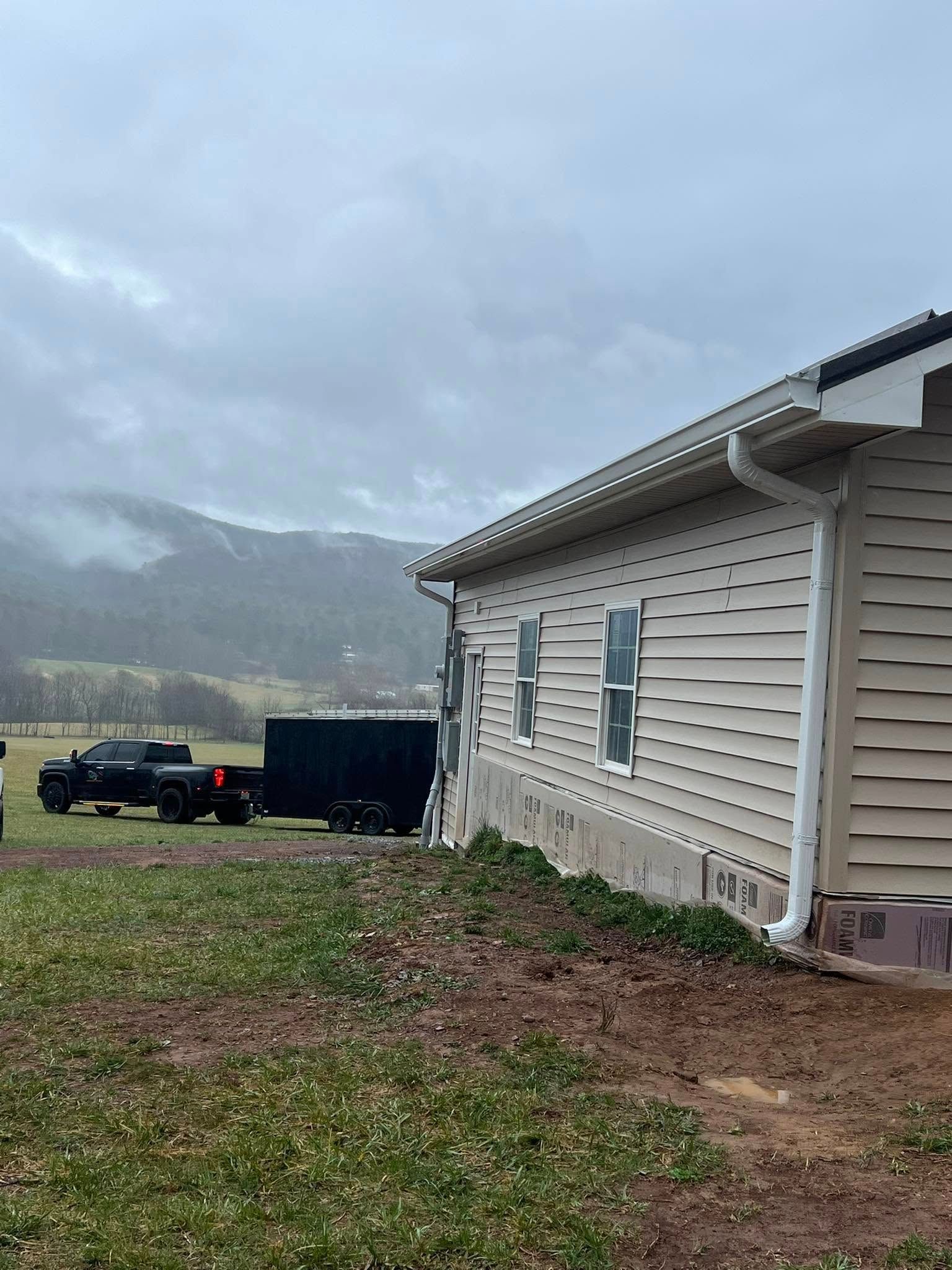 A side view of a beige house with a black trailer and truck parked on a grass field before misty, mountainous hills.