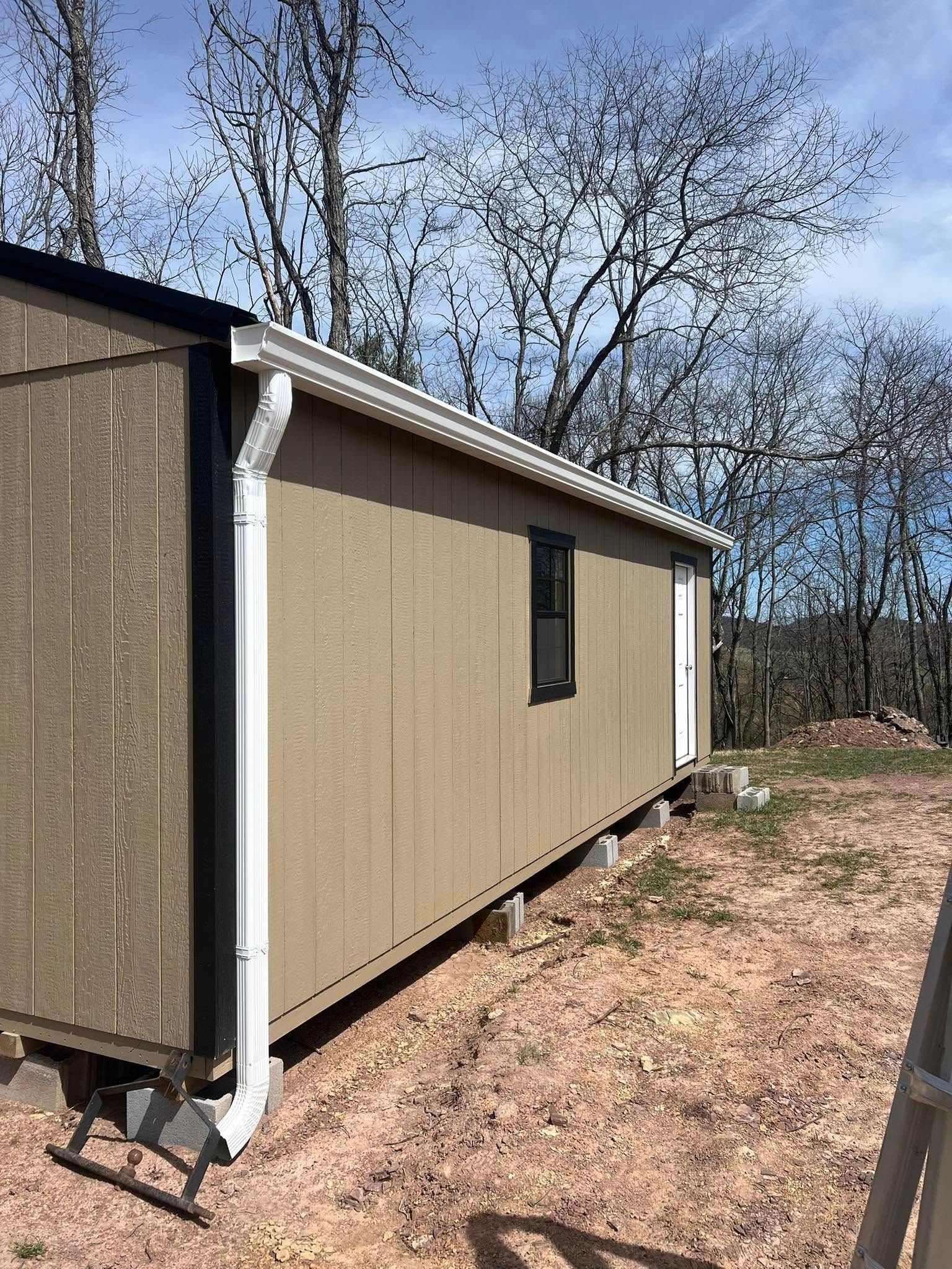 Tan shed with a black door frame and white gutters, sitting on concrete block supports in a wooded, dirt clearing.