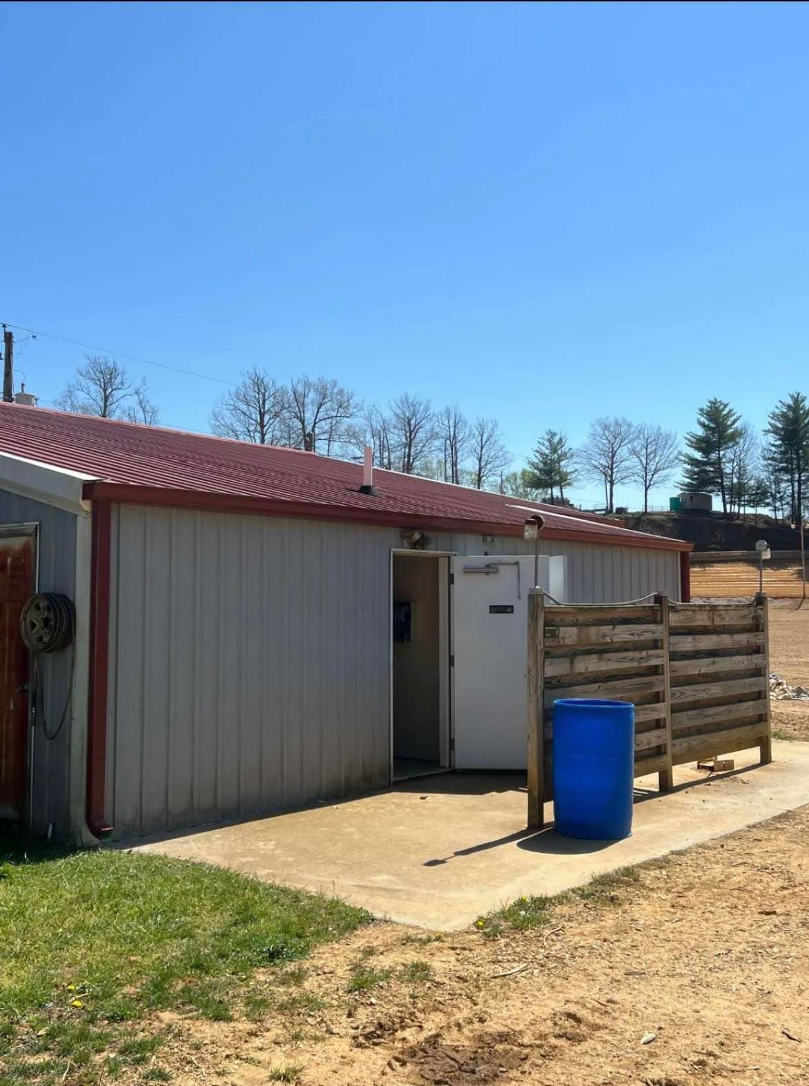 A light gray metal building with a maroon roof, an open doorway, and a blue plastic barrel on a concrete pad.