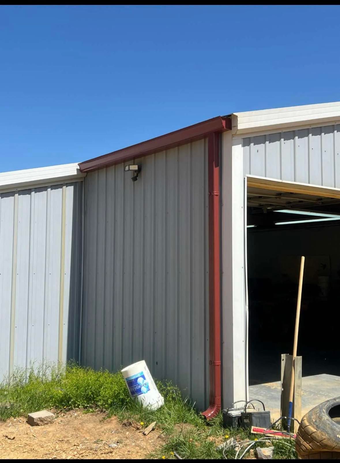 A corner view of a grey metal workshop exterior with a red trim, a white bucket on the ground, and a clear blue sky.
