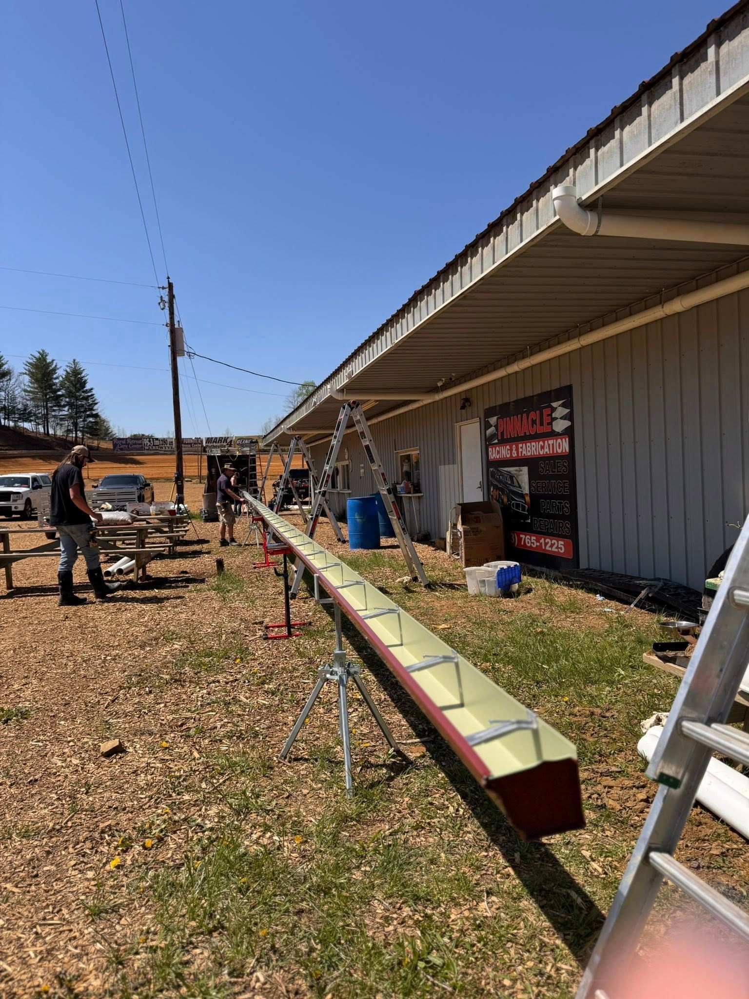 A long, newly fabricated metal gutter rests on tripods outdoors, with a worker standing nearby beside a building.