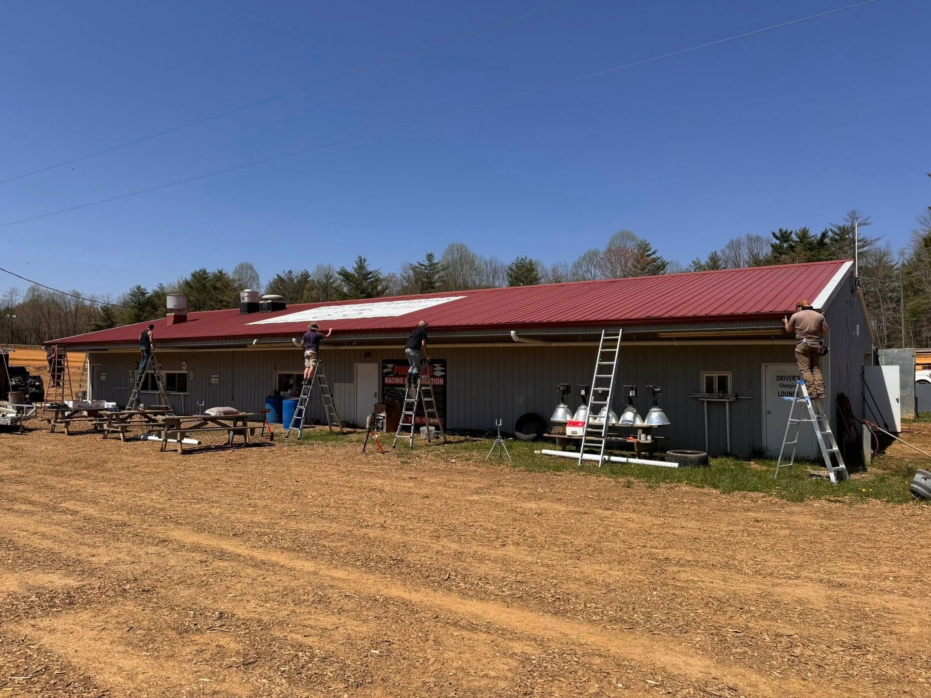 Workers on ladders paint the metal roof of a single-story building on a sunny day with a dirt yard.
