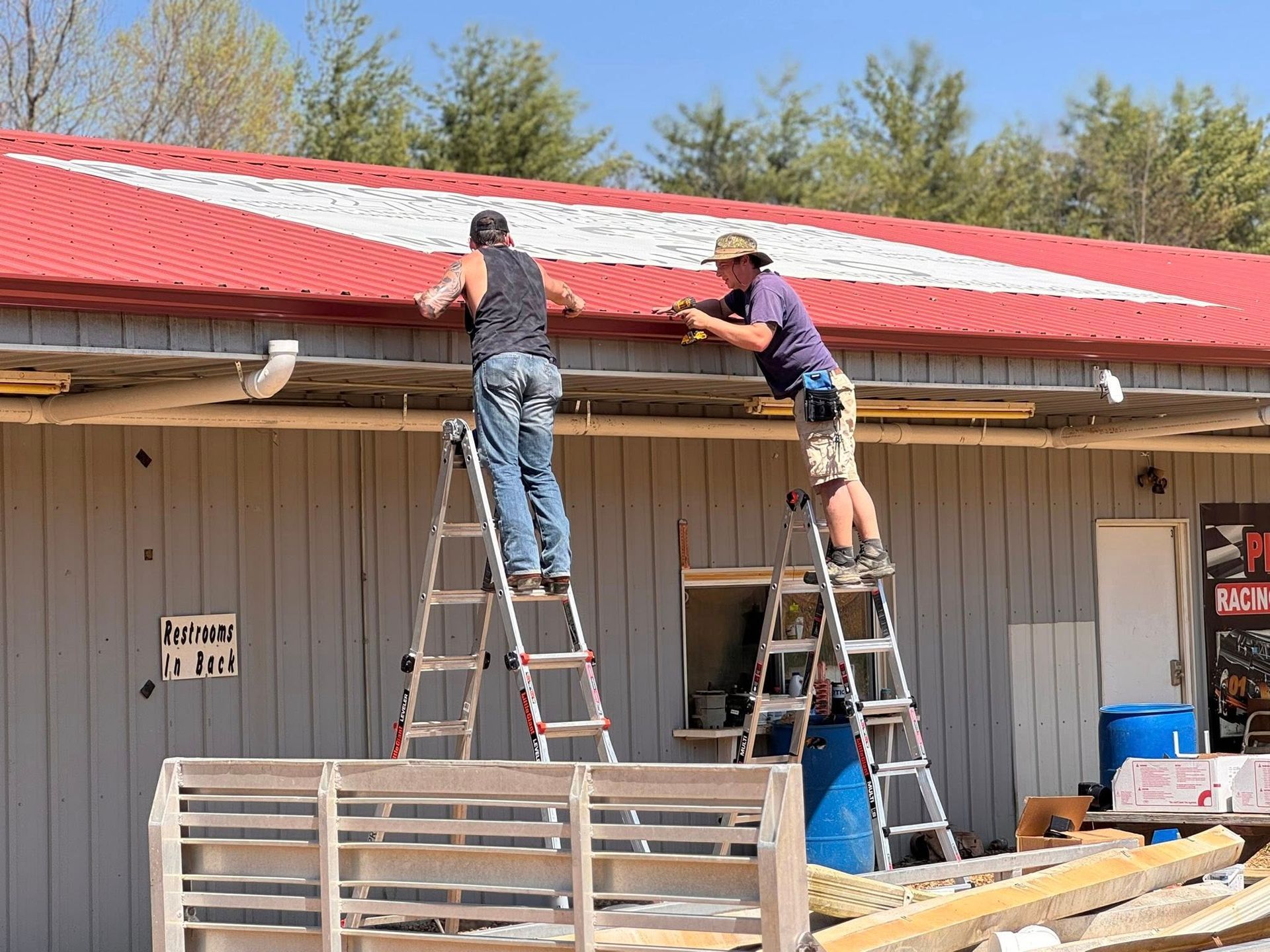 Two people stand on ladders, installing trim on the edge of a building with a red metal roof under a bright blue sky.
