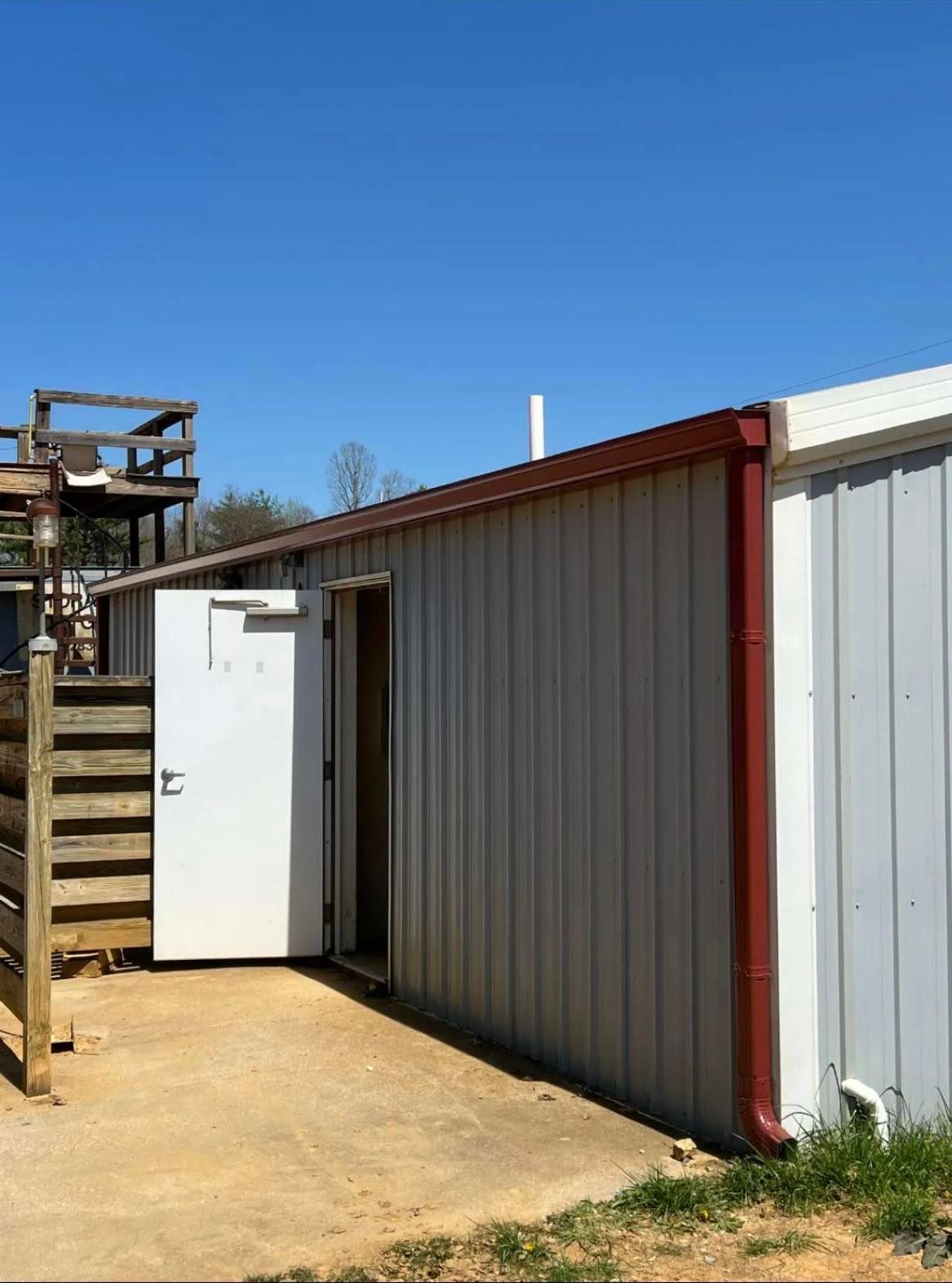 A metal storage building under a clear blue sky, featuring a white door left open and a dark red trim along the roofline.