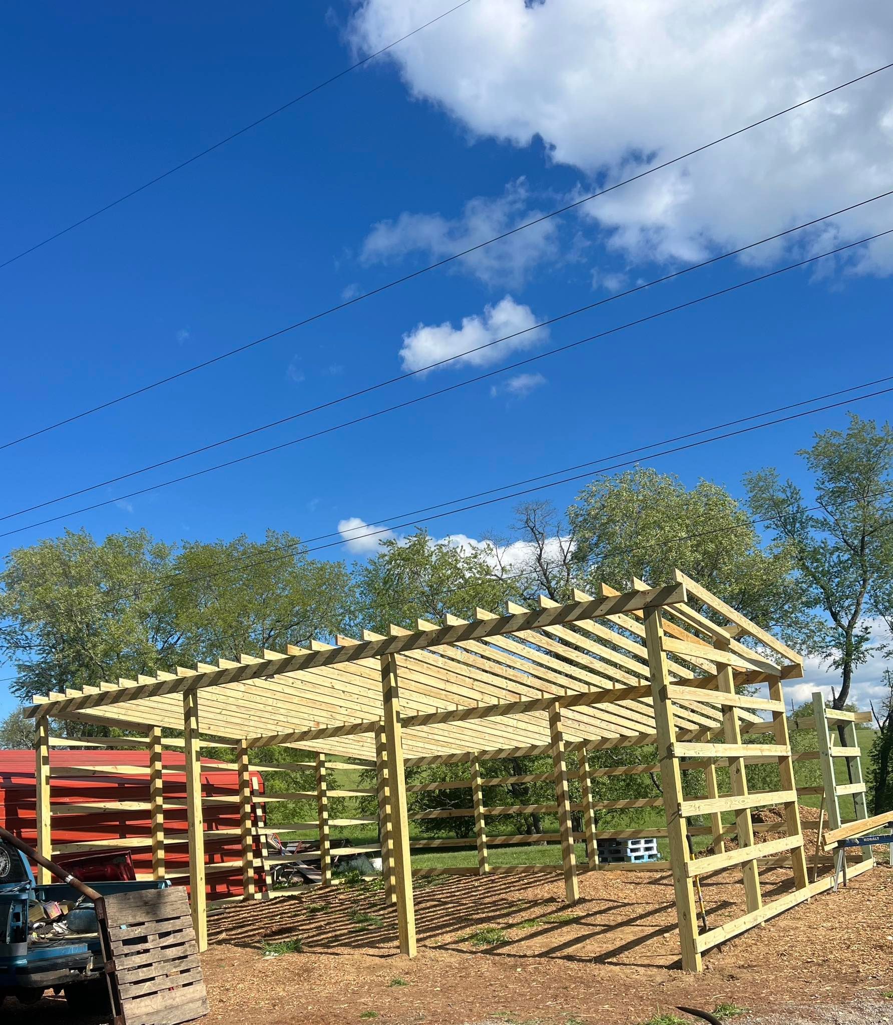 A large, wooden pergola structure stands in an outdoor area under a clear, sunny blue sky with trees in the background.