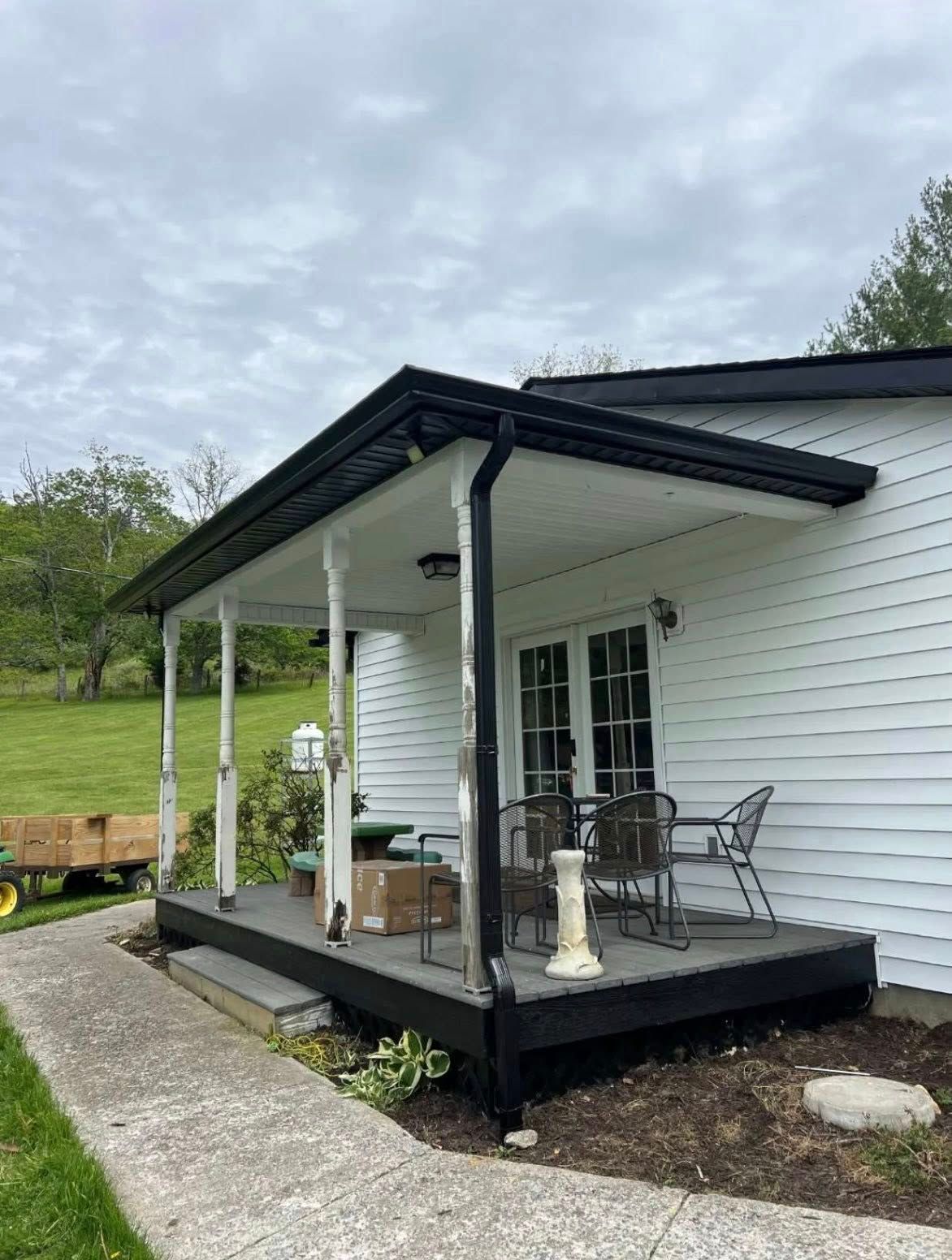 A white house with a covered wooden porch, black trim, and outdoor furniture, set against a green, grassy landscape.