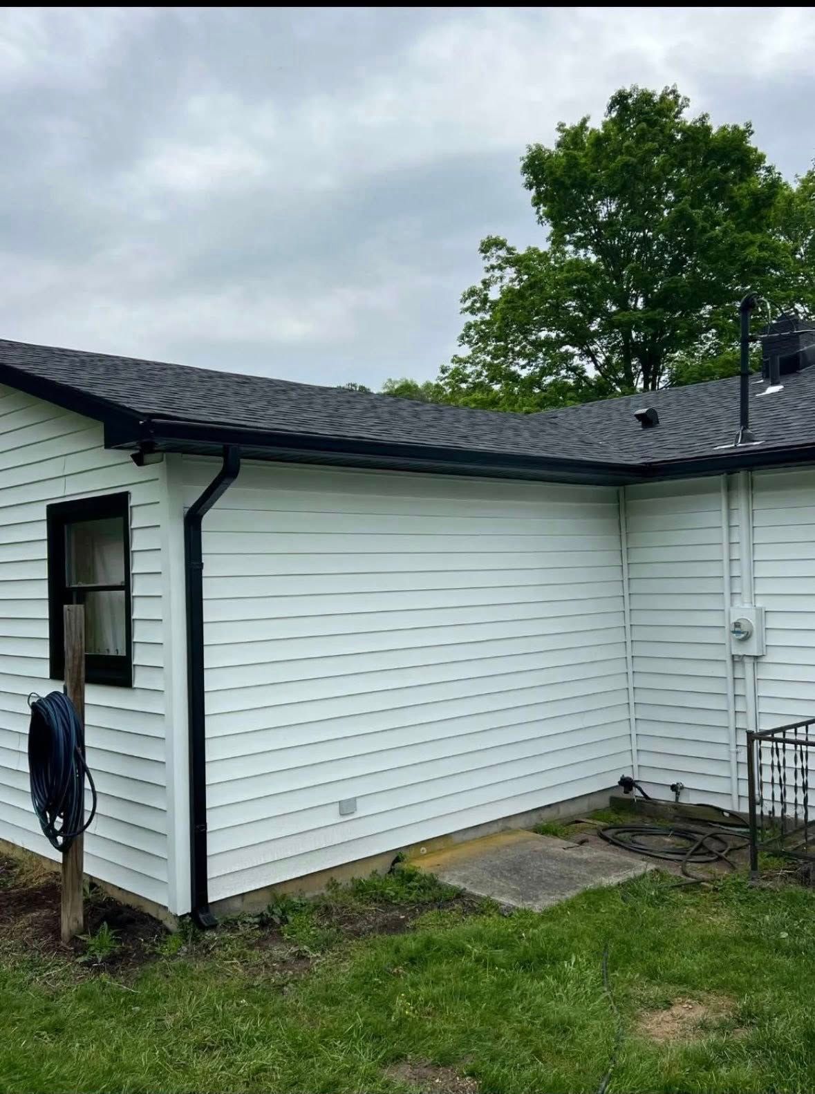 A corner view of a white horizontal-sided house with a black roof, black window trim, and black gutters against a cloudy sky.