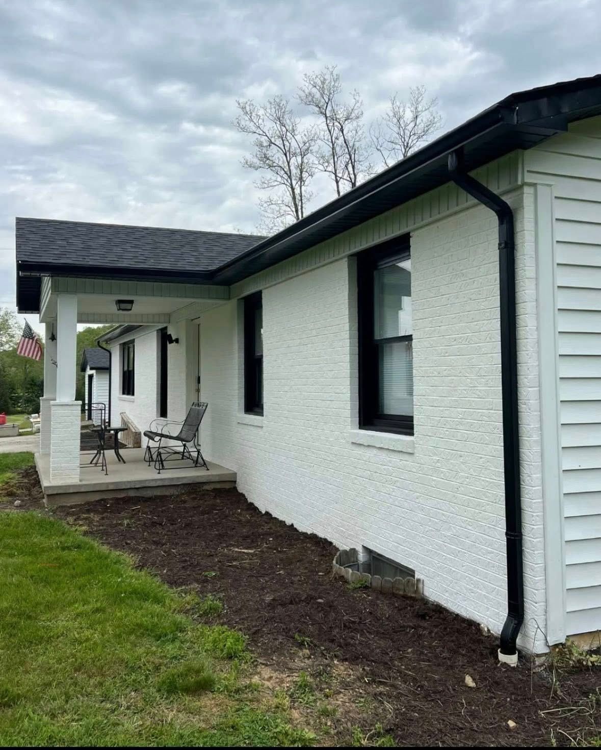 A white brick house exterior with black trim, a covered porch with seating, and dark mulch landscaping.