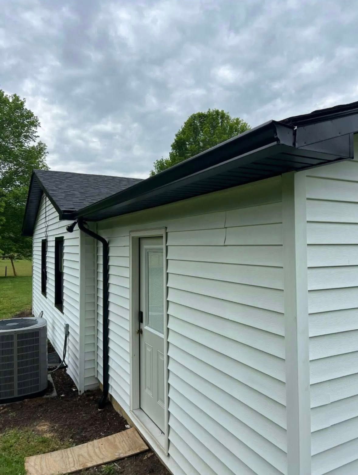 Side view of a white house exterior with black roof shingles, gutters, and window trim, next to an outdoor AC unit.
