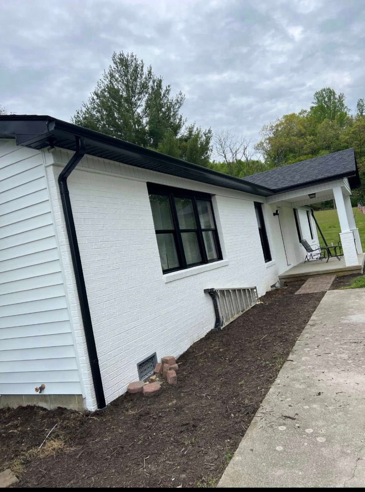 A white house with a black roof and black window frames, featuring a side porch and a mulched garden bed.
