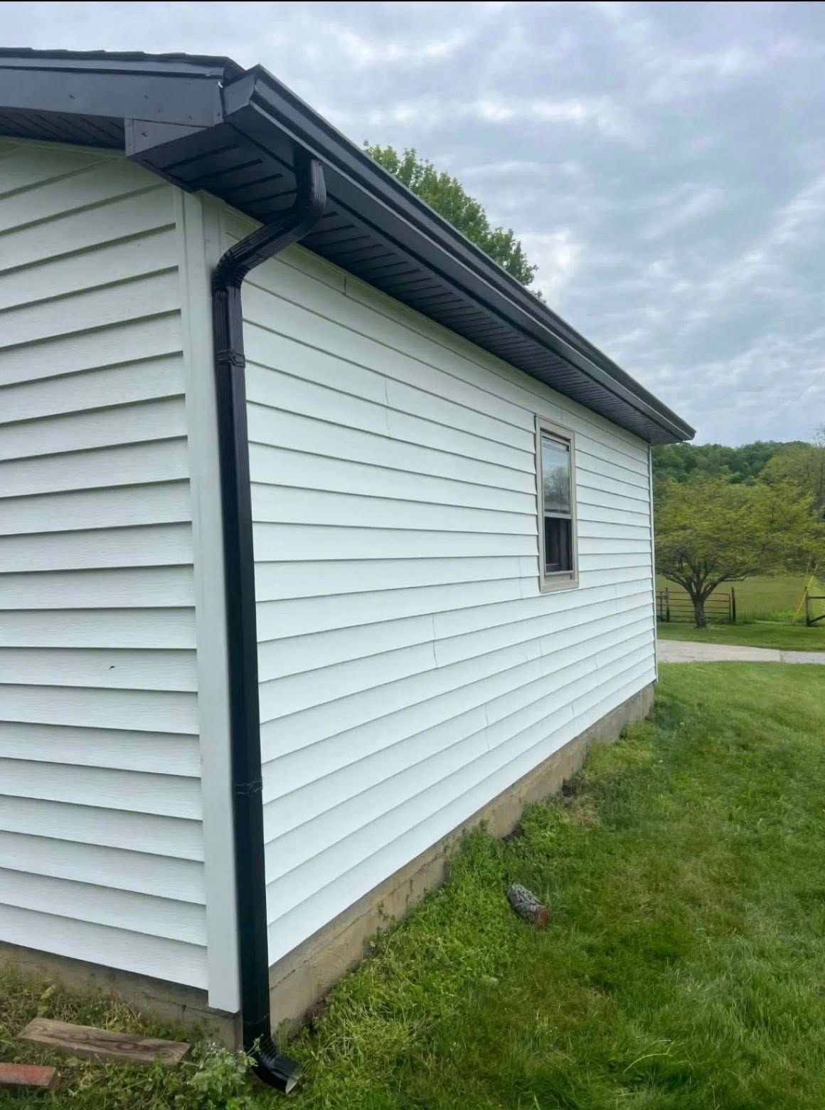 A side view of a white-sided house with a black gutter system and downspout, set against a green lawn and cloudy sky.