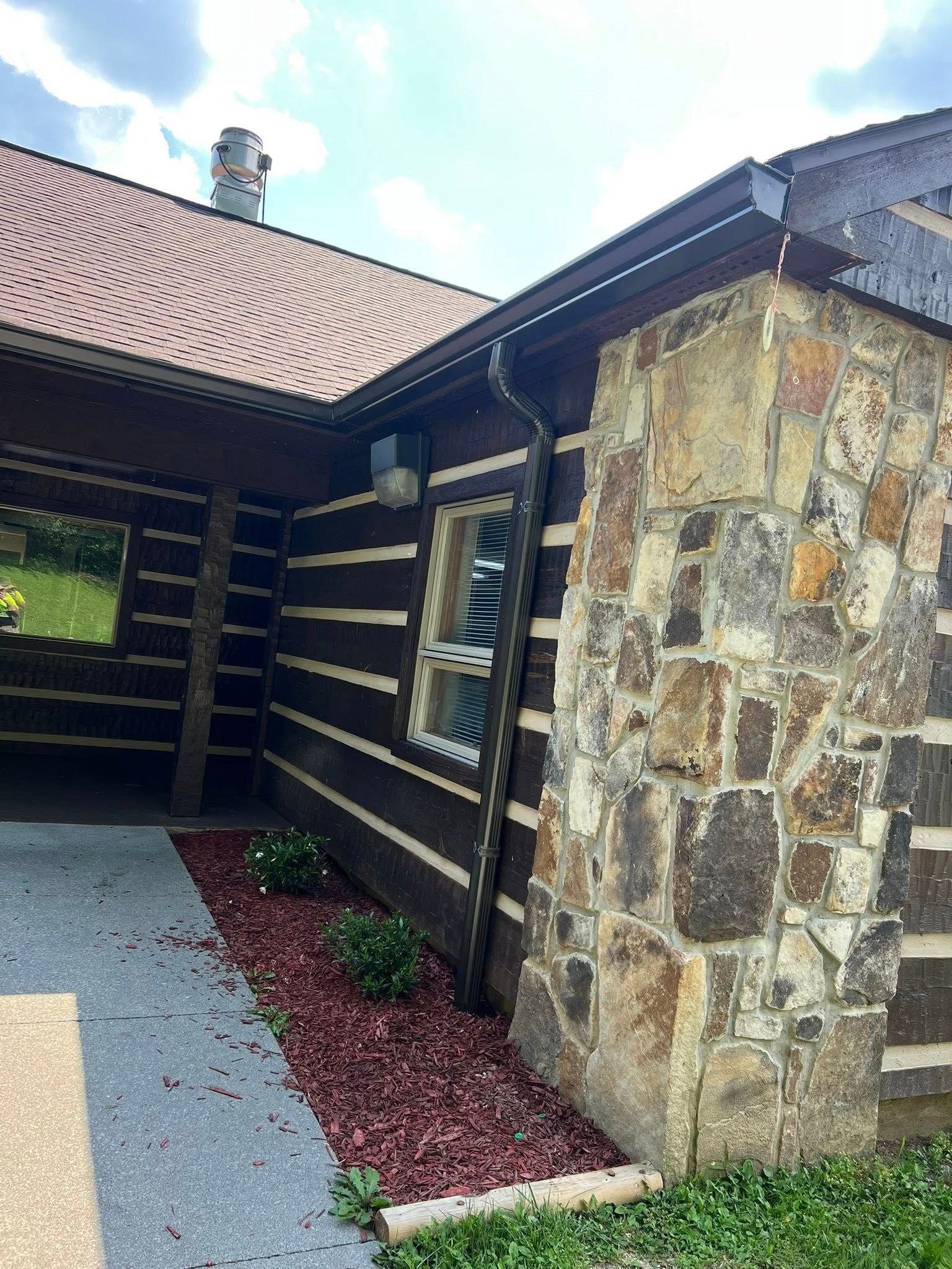 A rustic cabin exterior featuring dark horizontal logs, a stone chimney, and a small flower bed with red mulch.