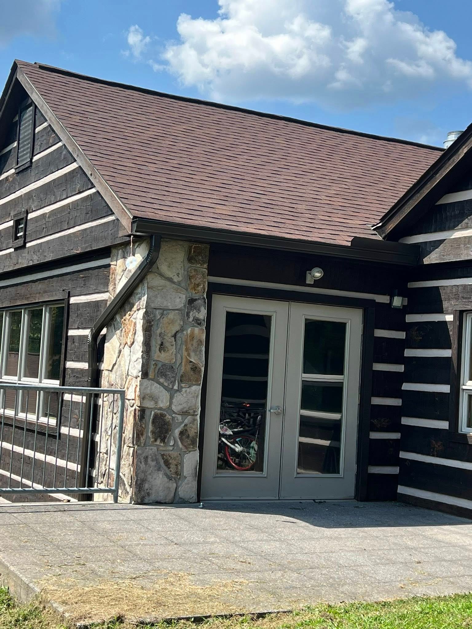 A log cabin building with a stone-accented entrance, double glass doors, a brown shingled roof, and a concrete patio.