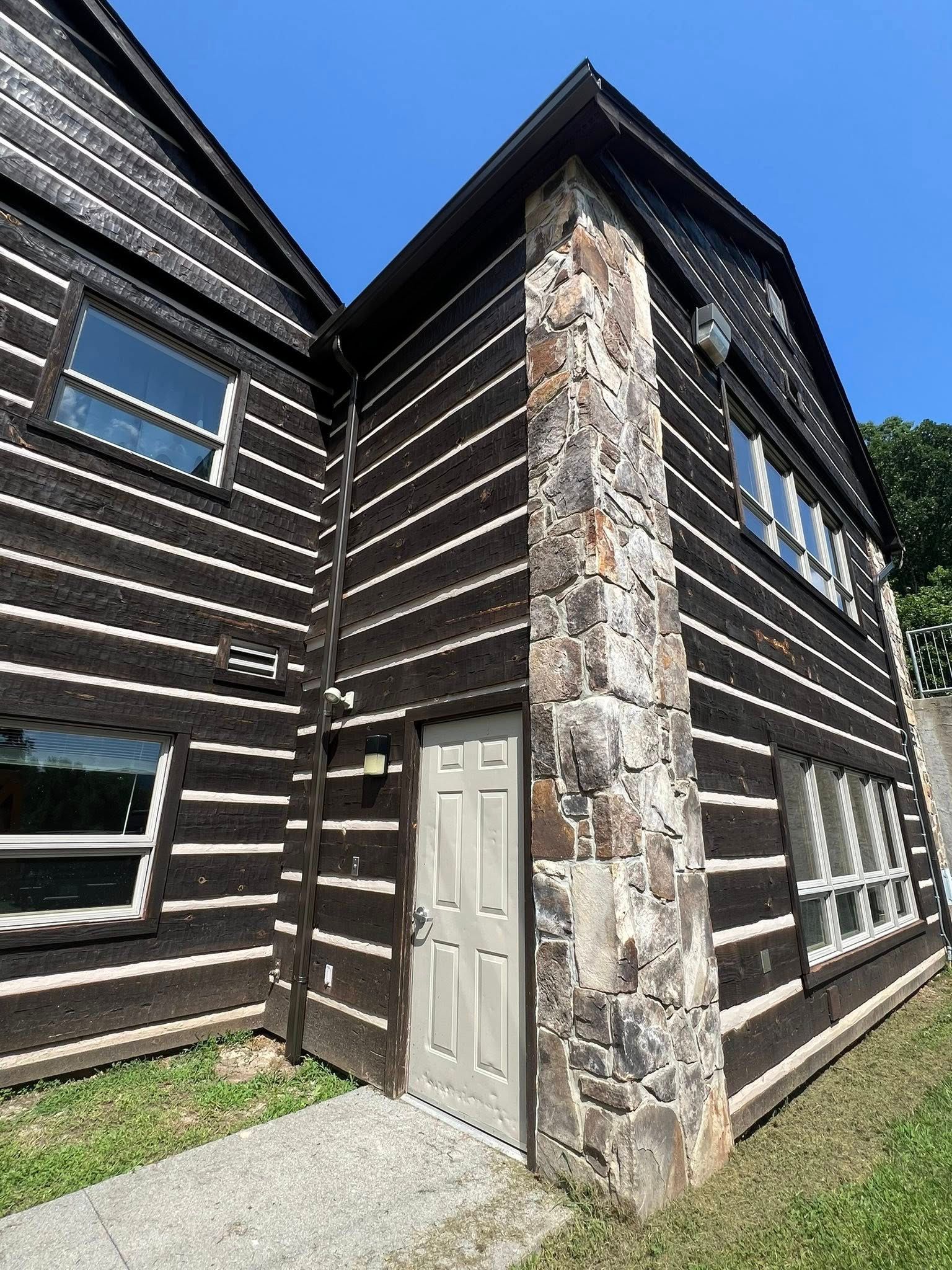 A two-story building with dark log-style horizontal siding, a central stone chimney, and a white door under a blue sky.