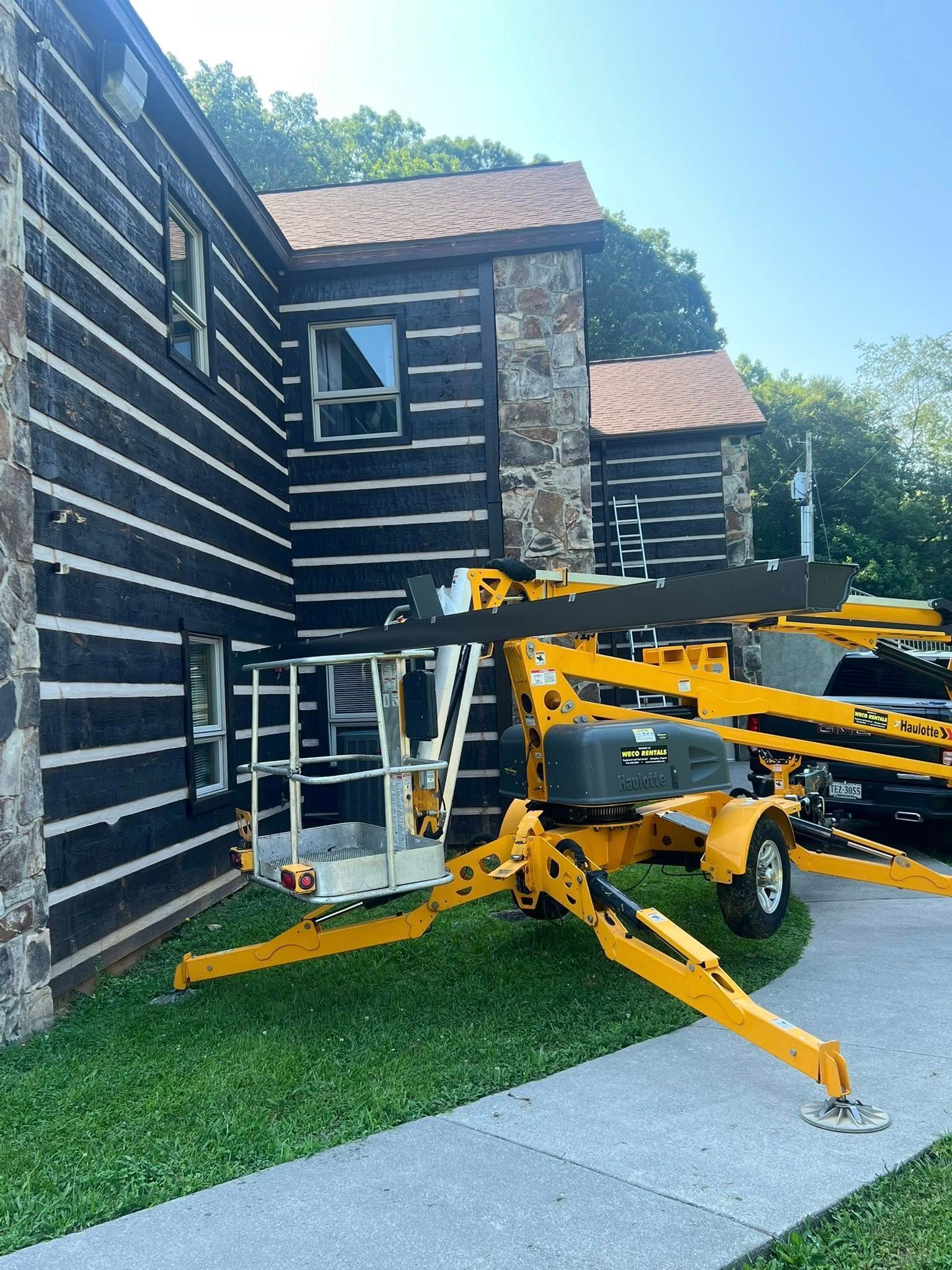 A bright yellow towable boom lift parked on grass next to the dark exterior walls of a rustic log and stone building.