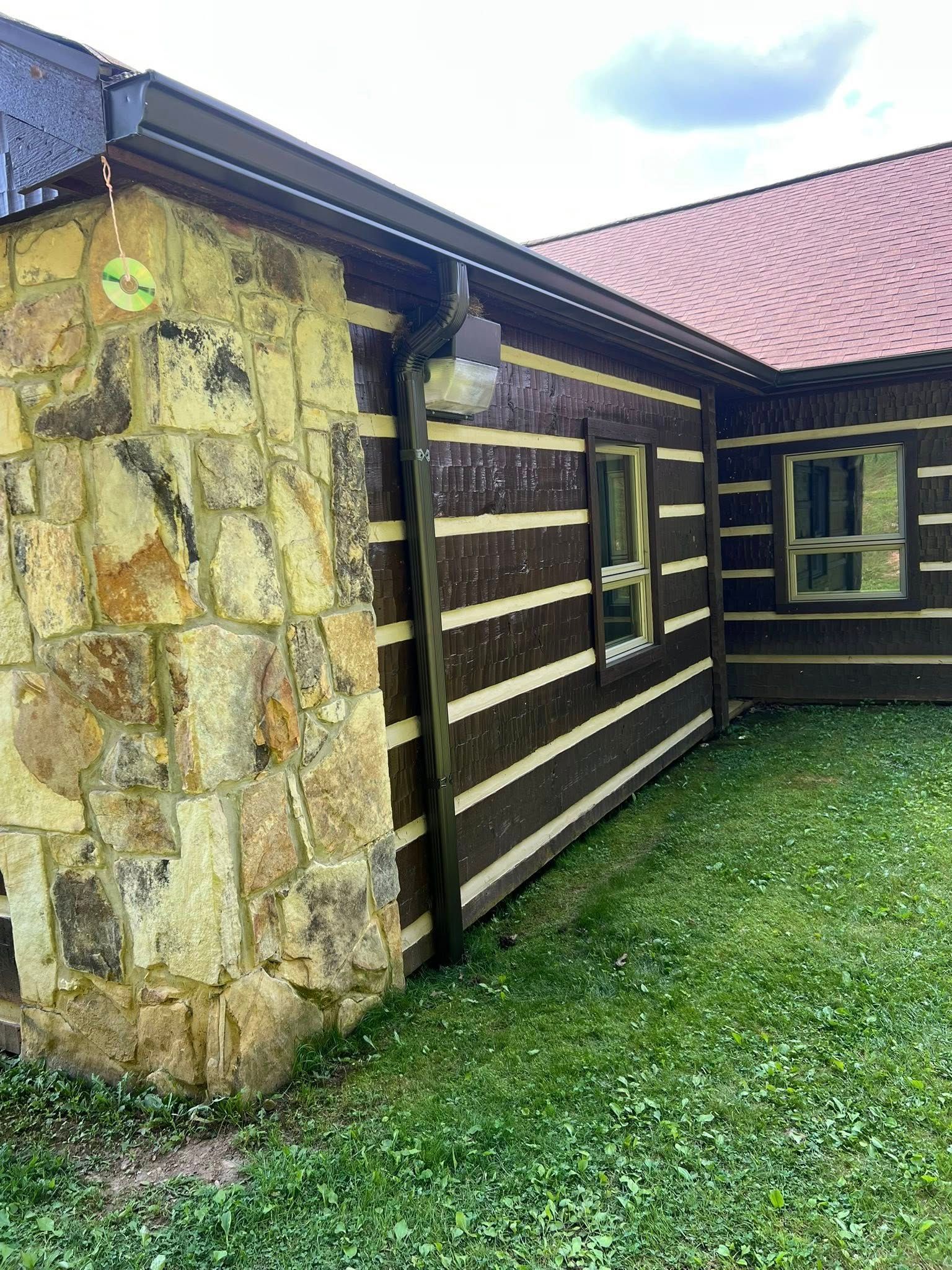A corner view of a rustic log cabin with a stone chimney and a red roof, set against a green grassy lawn.