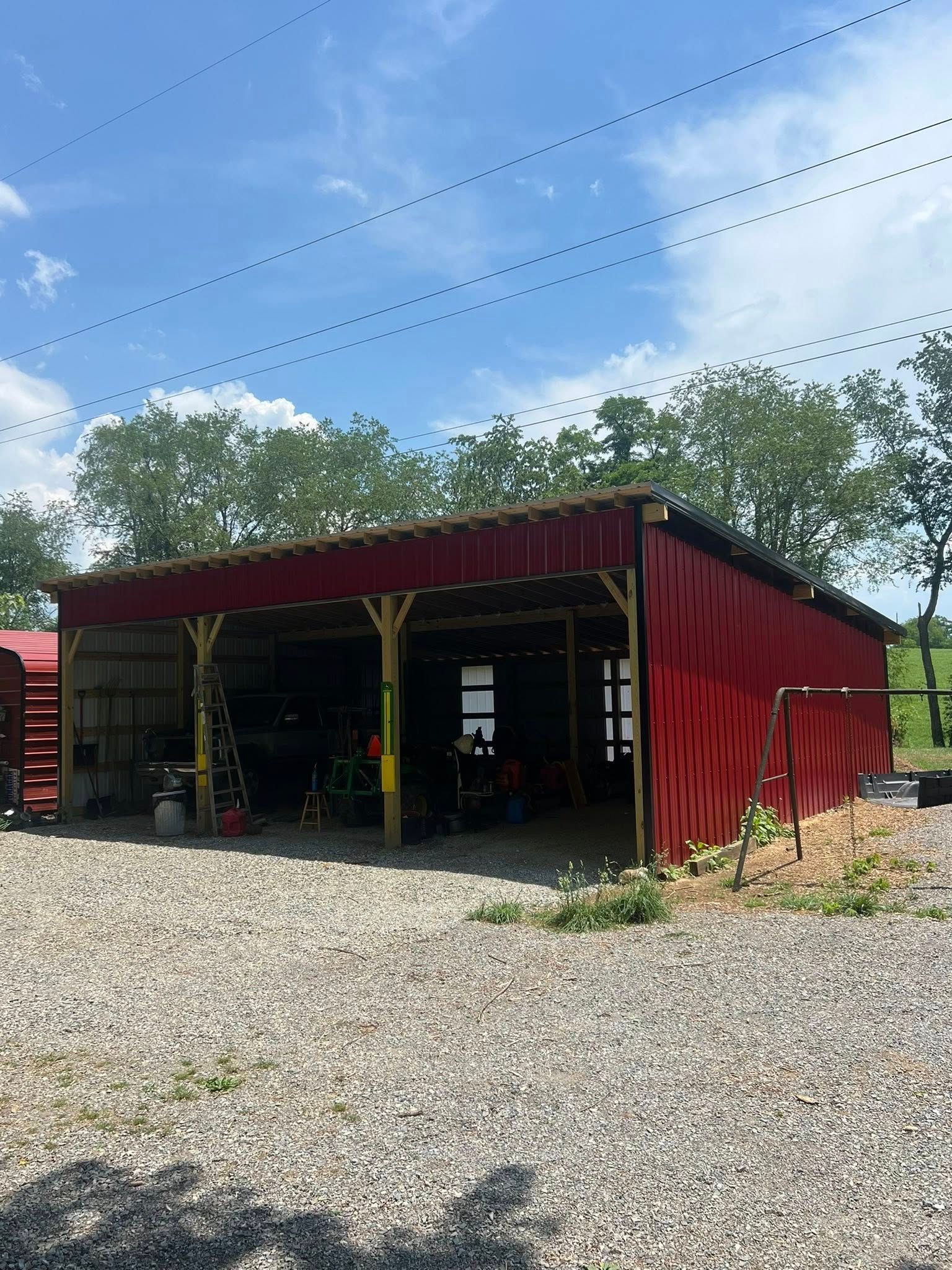 A red, open-sided pole barn structure sits on a gravel lot under a bright blue sky with power lines overhead.