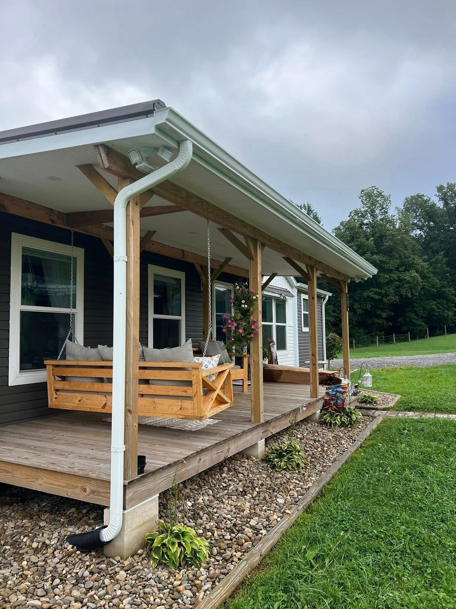 A wooden porch with a pallet swing, white trim, and a downspout, set against a dark house and a grassy, gravel yard.
