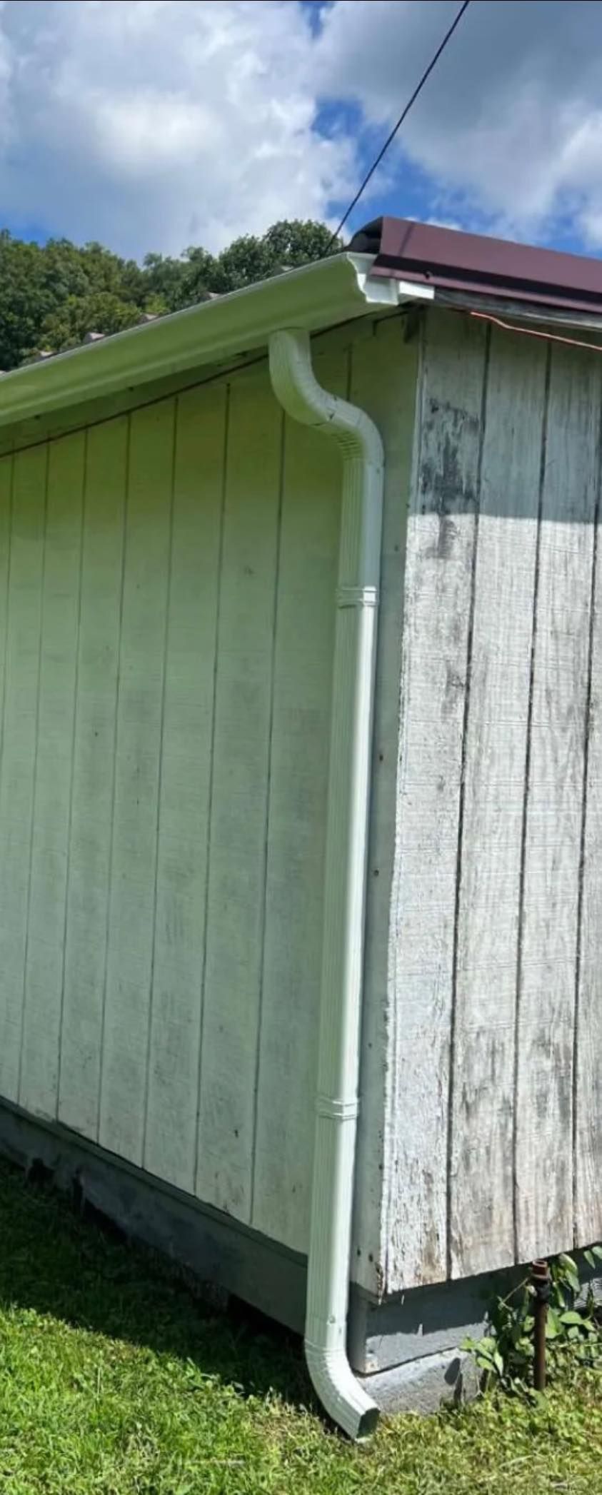 A white gutter downspout attached to the corner of a building with light green and white weathered wood siding.