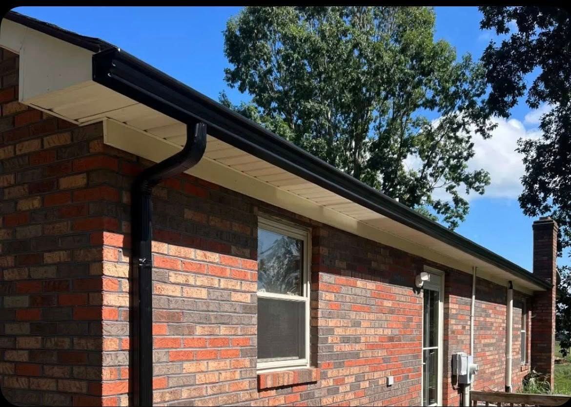 Exterior view of a brick house featuring a dark gutter and downspout, white soffit, and a window against a blue sky.