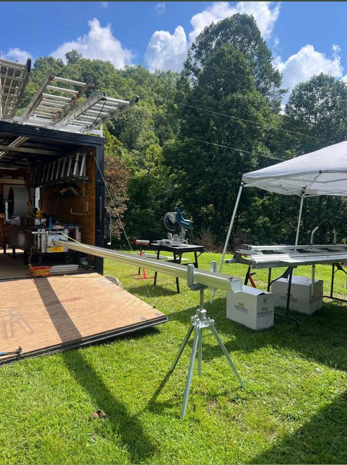 A work trailer and a portable canopy set up on a lawn, with a metal siding brake mounted on a tripod in the foreground.