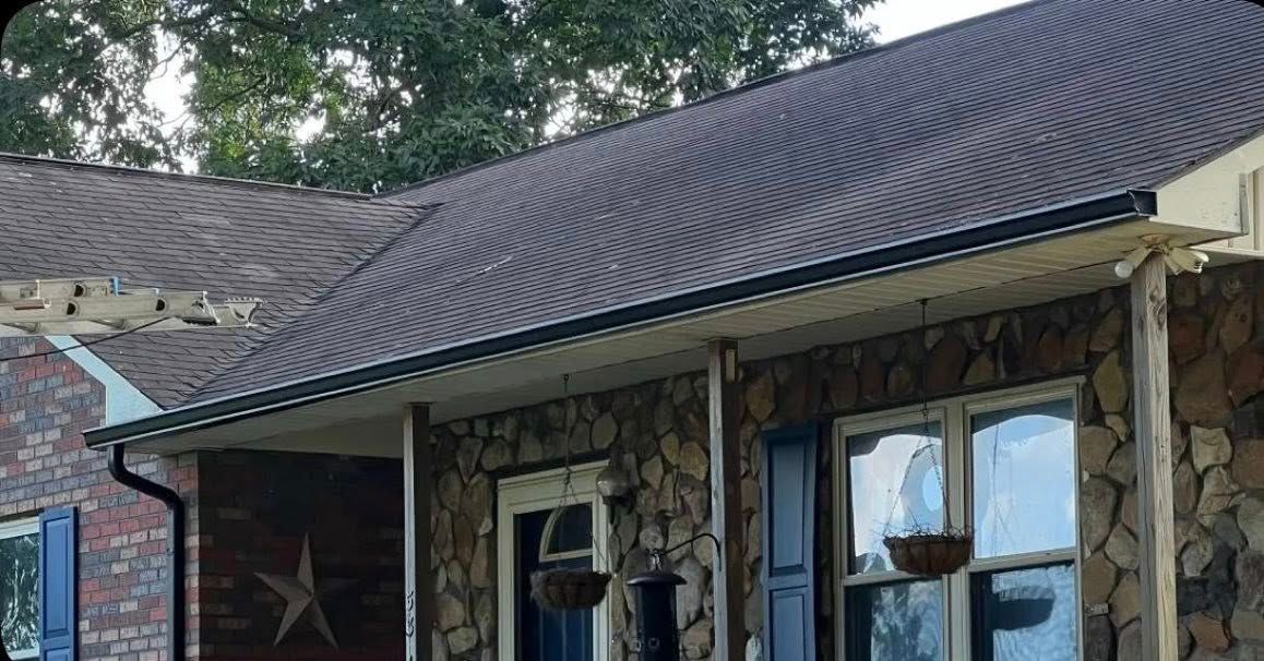 A residential stone-front house with a dark shingled roof, blue shutters, and a covered porch on a sunny day.
