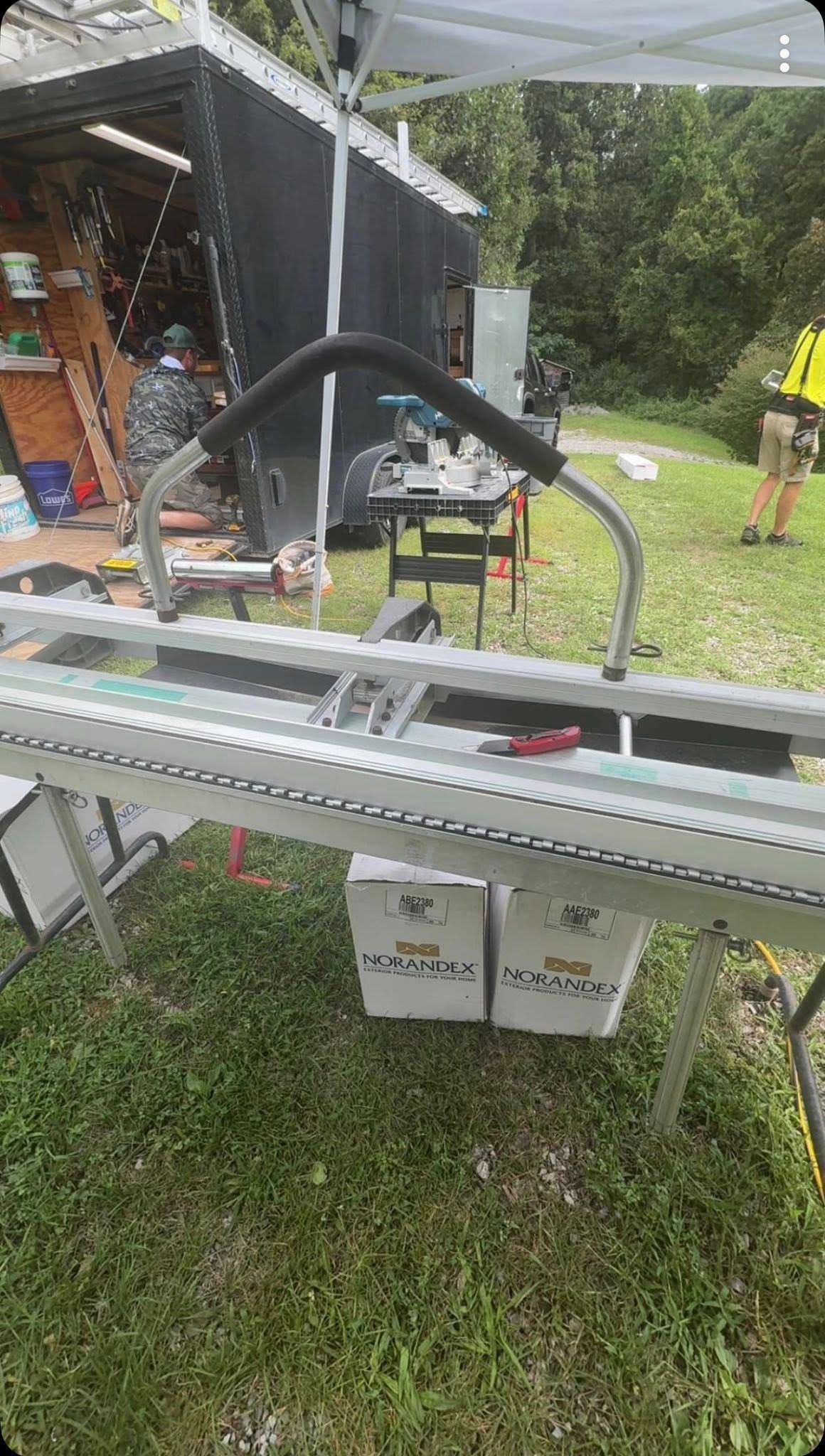 A metal siding brake sits on a grassy field in front of a trailer, with a person standing nearby in the background.