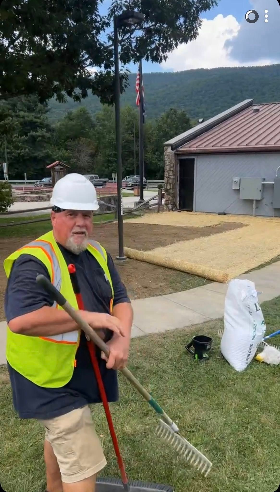 A person in a safety vest and hard hat holds a rake while standing on a lawn next to a building and landscaping materials.